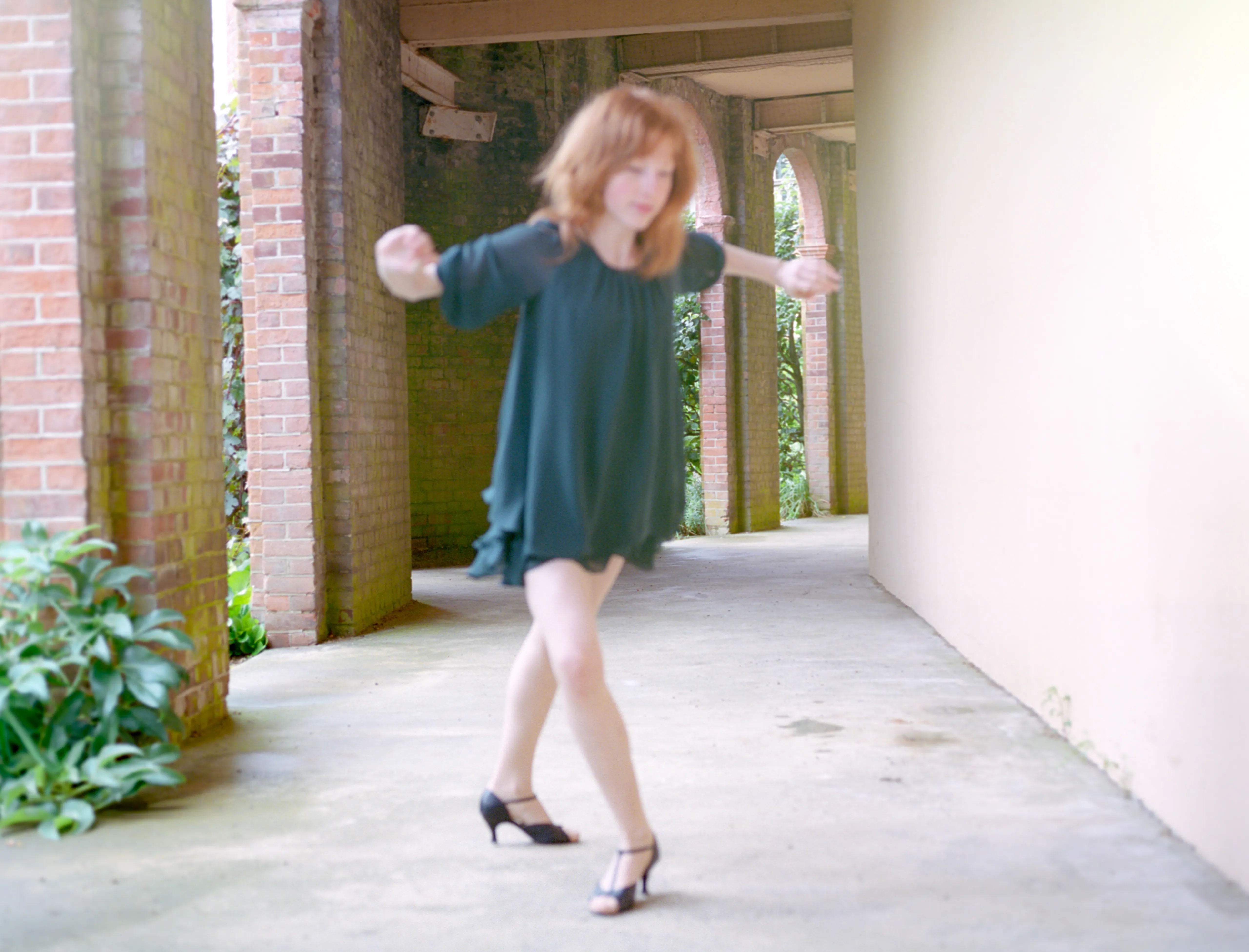 Clélia spins in a teal chiffon dress in a brick colonnade, arms out and hem flaring.