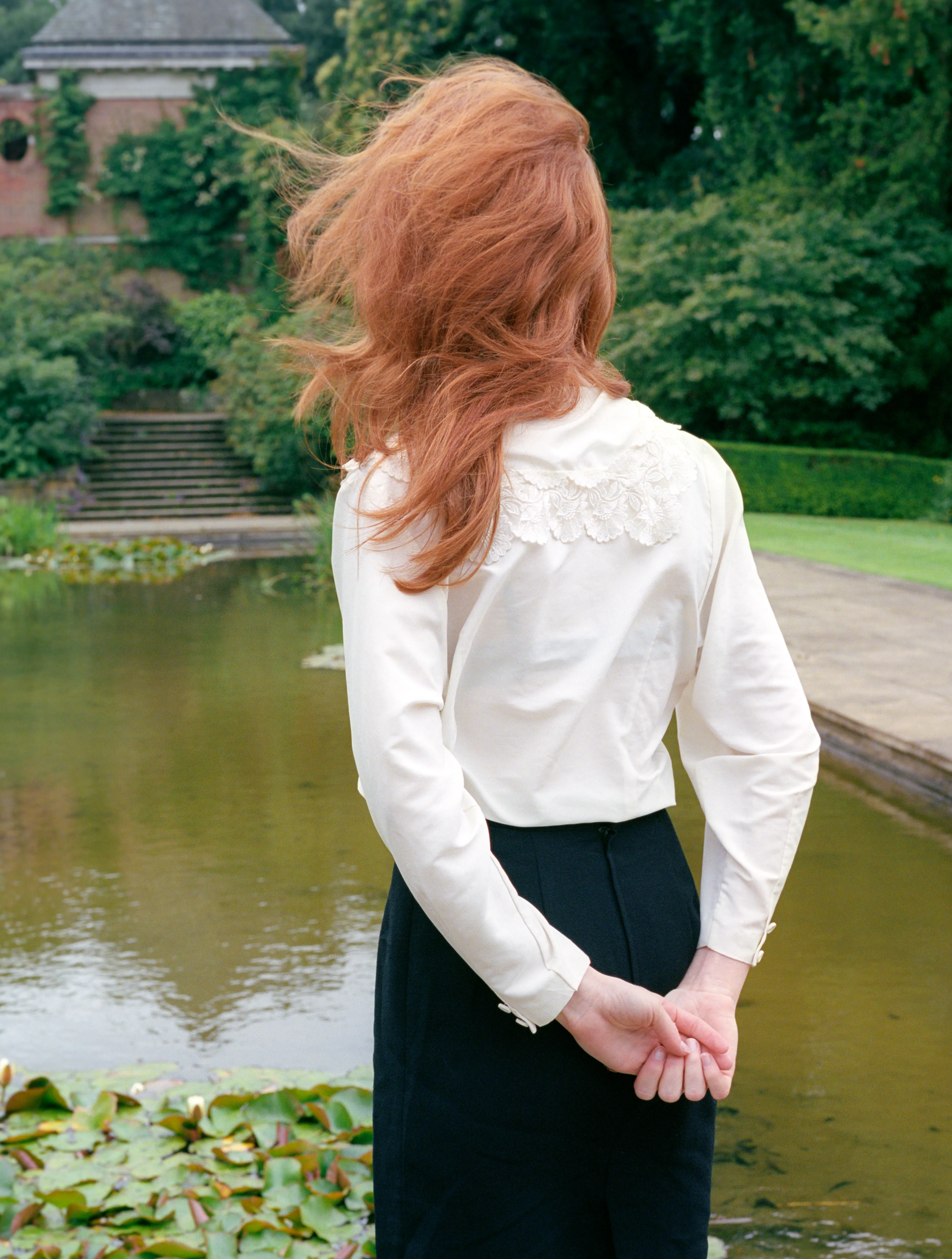 Seen from behind, Clélia stands by a garden pond in a white blouse, her hair swept across her face.
