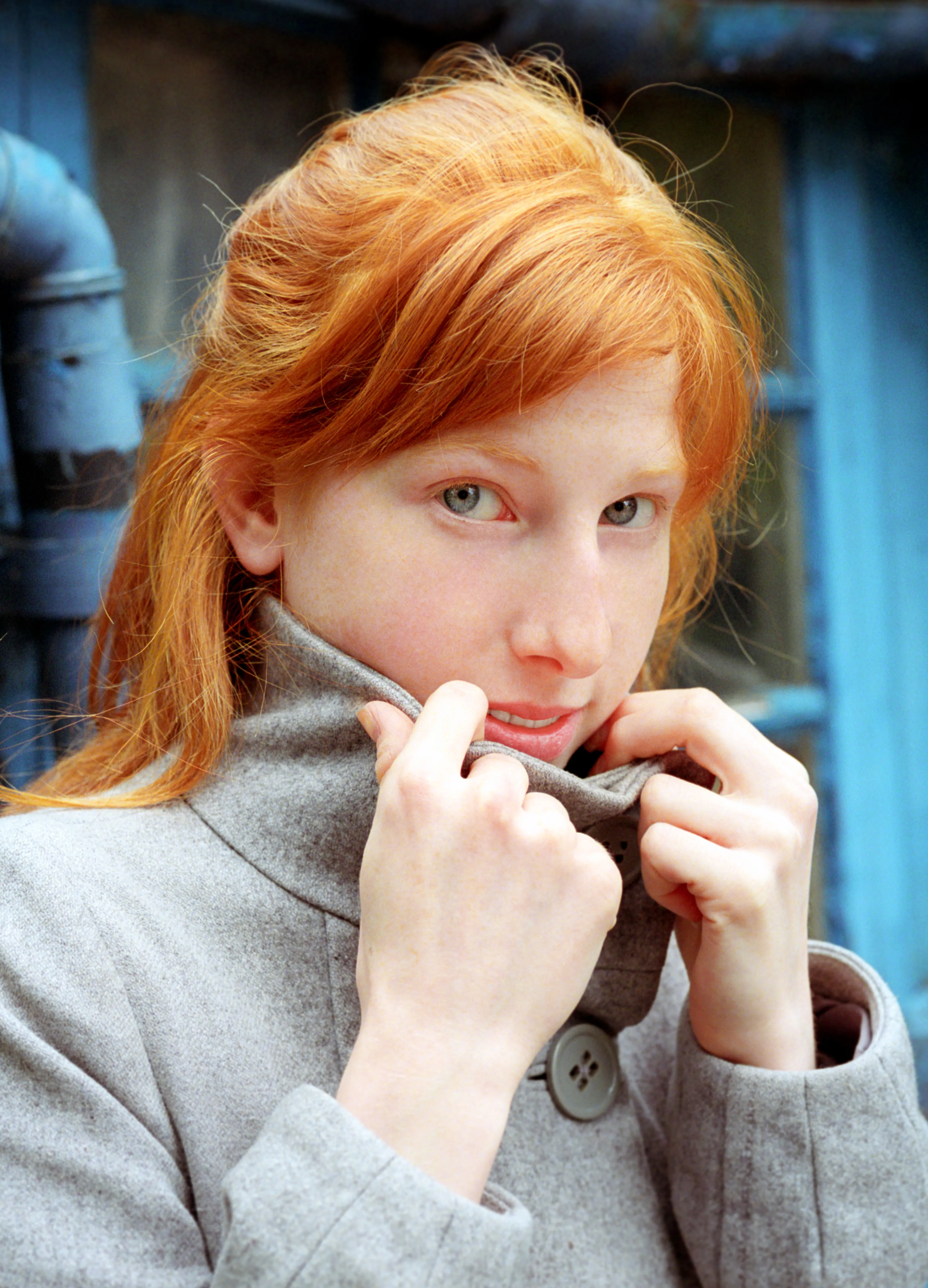 Clélia pulls up the collar of her grey coat with both hands, against blue-painted ironwork, red hair loose.
