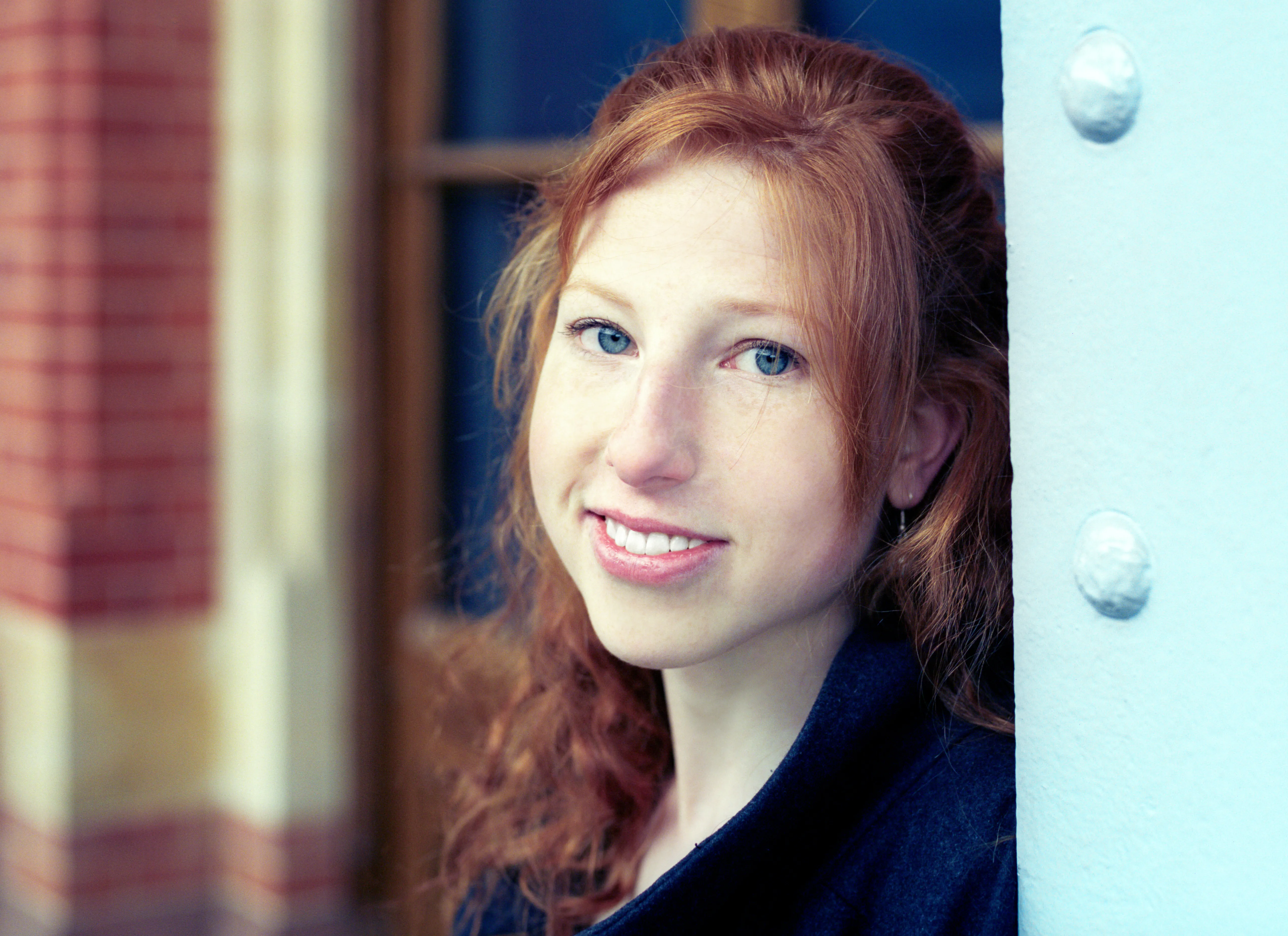 A close portrait of Clélia smiling, red hair down, beside a blue door and red brick.