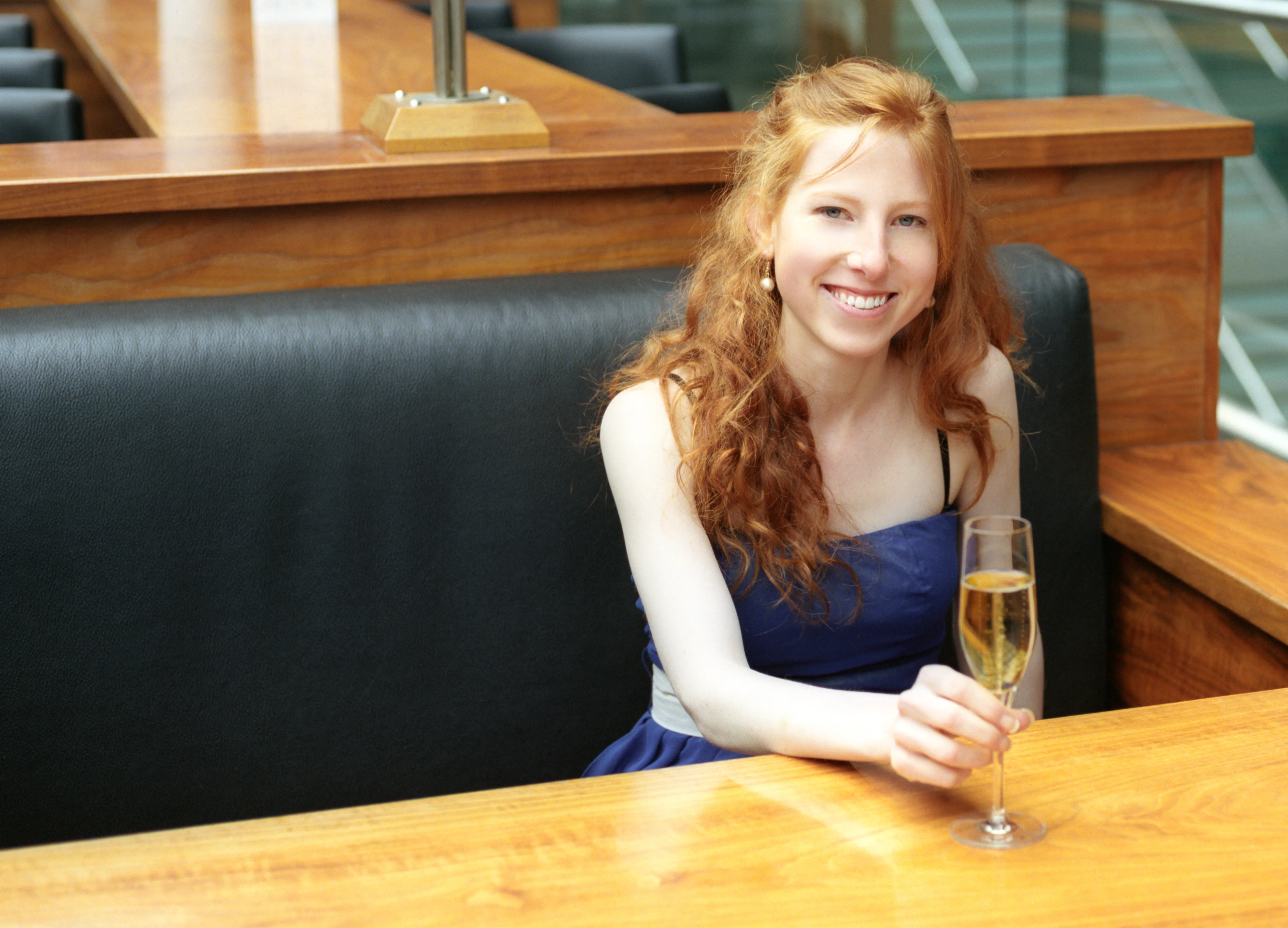 Clélia sits at a wooden bar with a champagne flute, smiling toward the camera, a dark leather banquette behind.