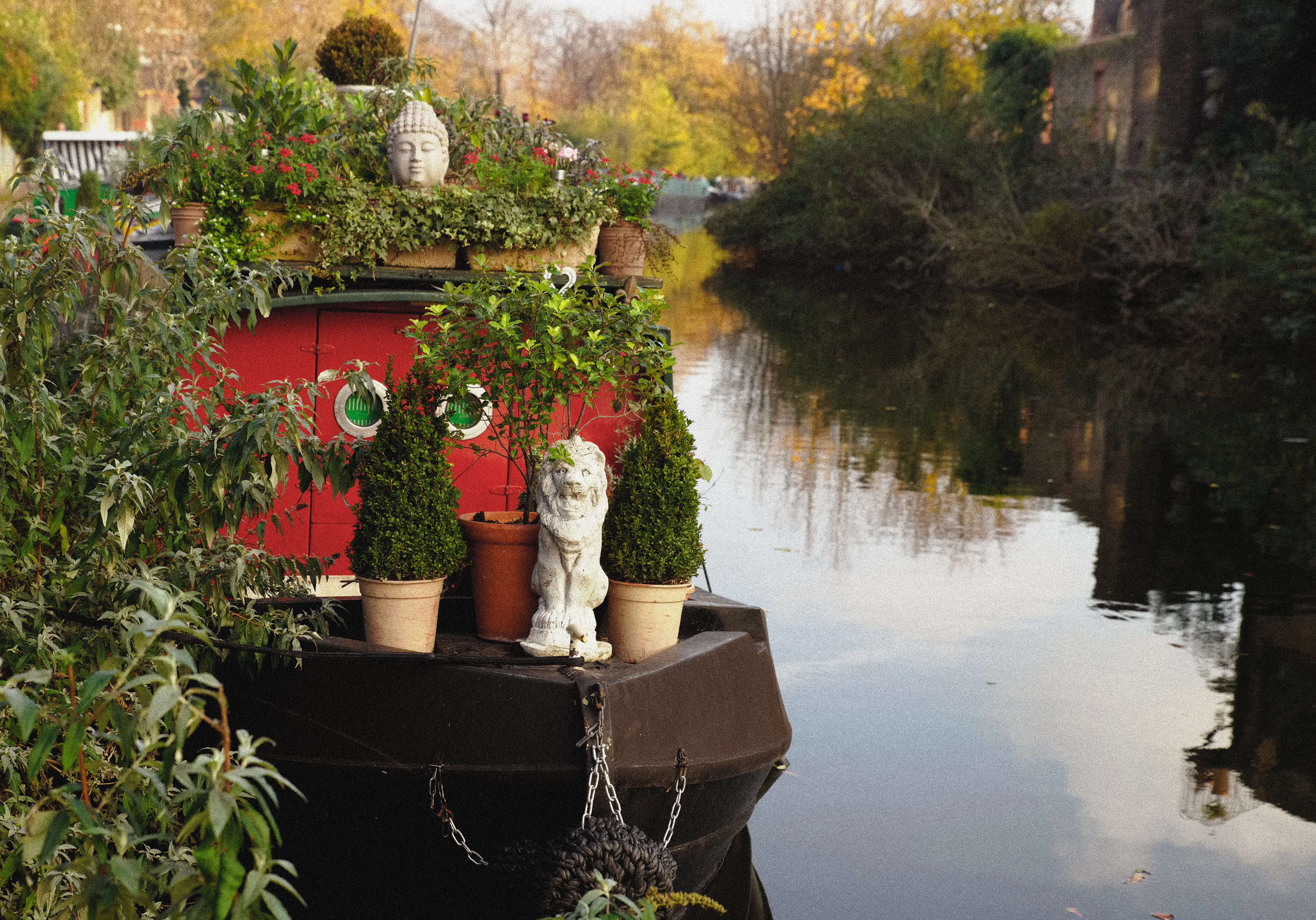 The decorated bow of a moored narrowboat on a still canal — potted bay trees, a stone lion and a Buddha head among the greenery.
