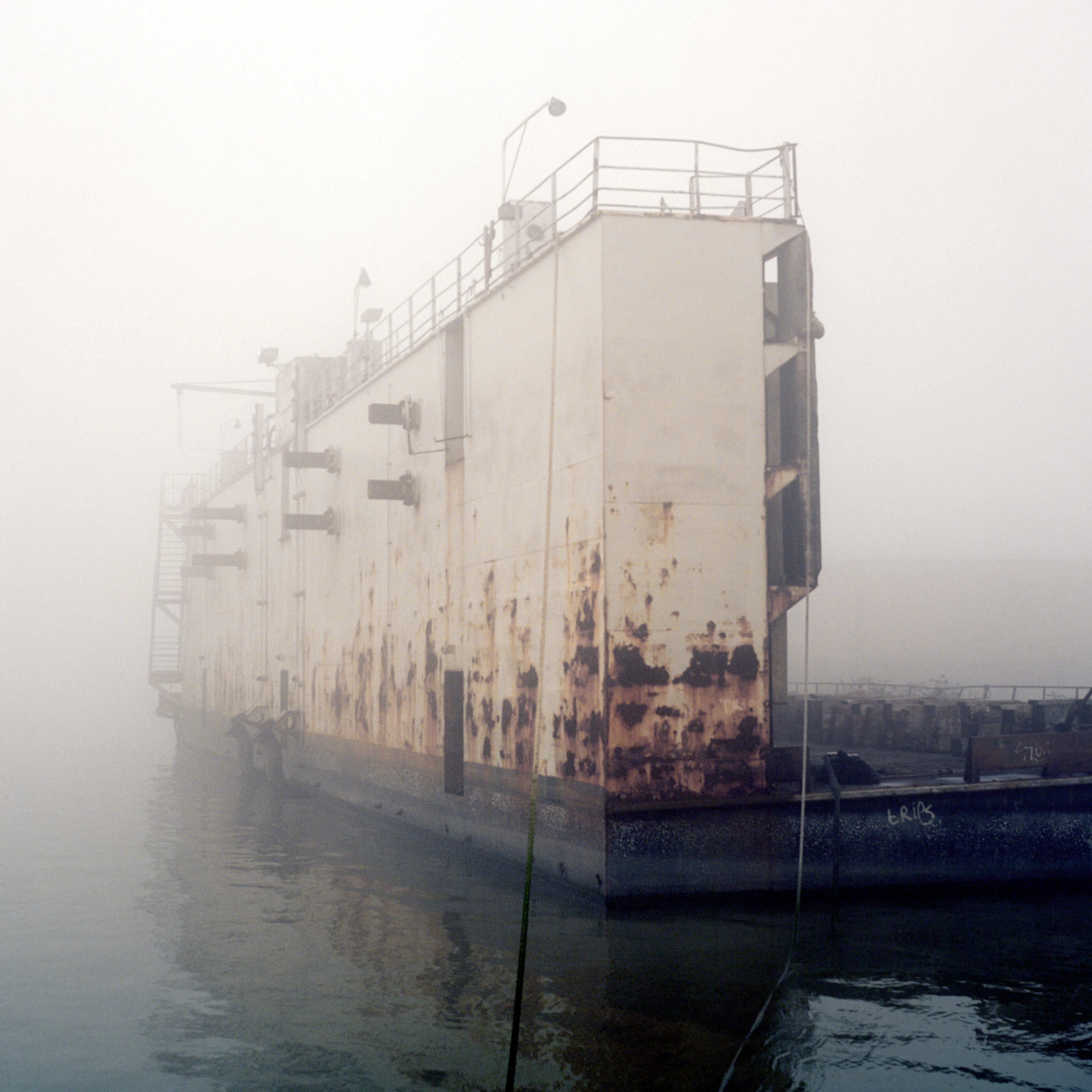 A rusted derelict barge moored in fog, its far end dissolving into white nothing over the water.