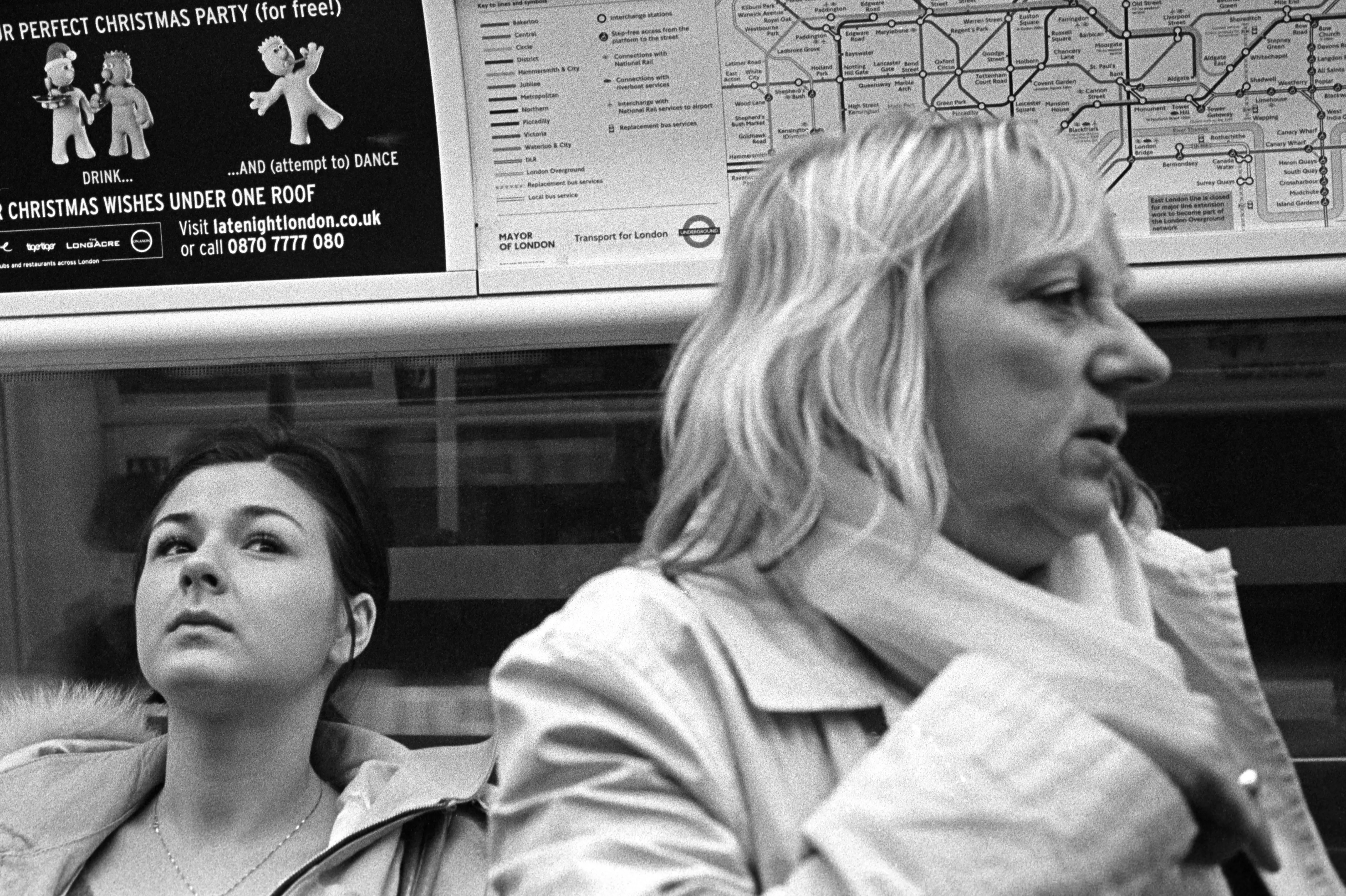 Two women in an Underground carriage beneath a Tube map and an advert — one seated and looking up, the other in profile.