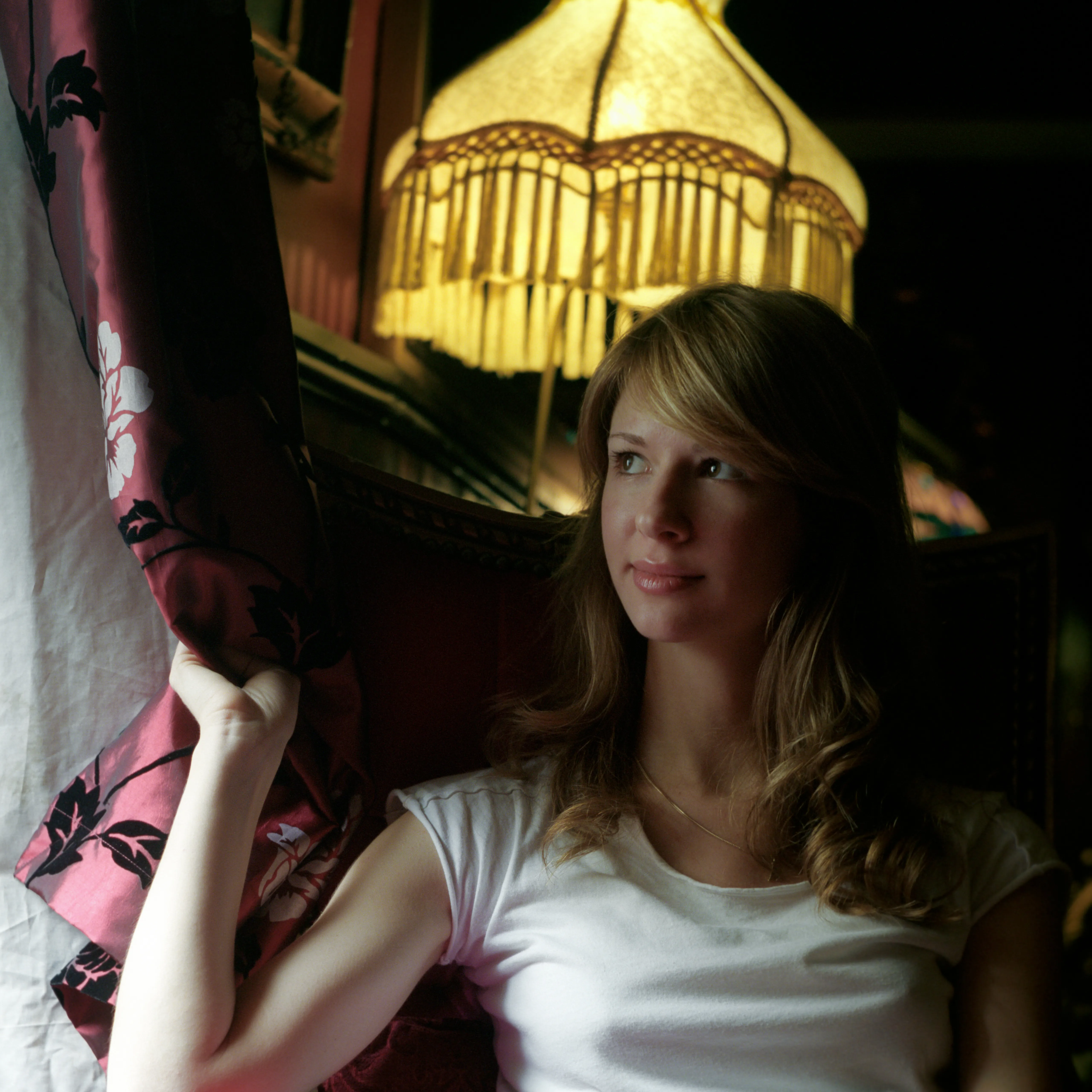Eva in lamplight, looking past the camera and holding back a dark floral curtain; a fringed lampshade glows above her.