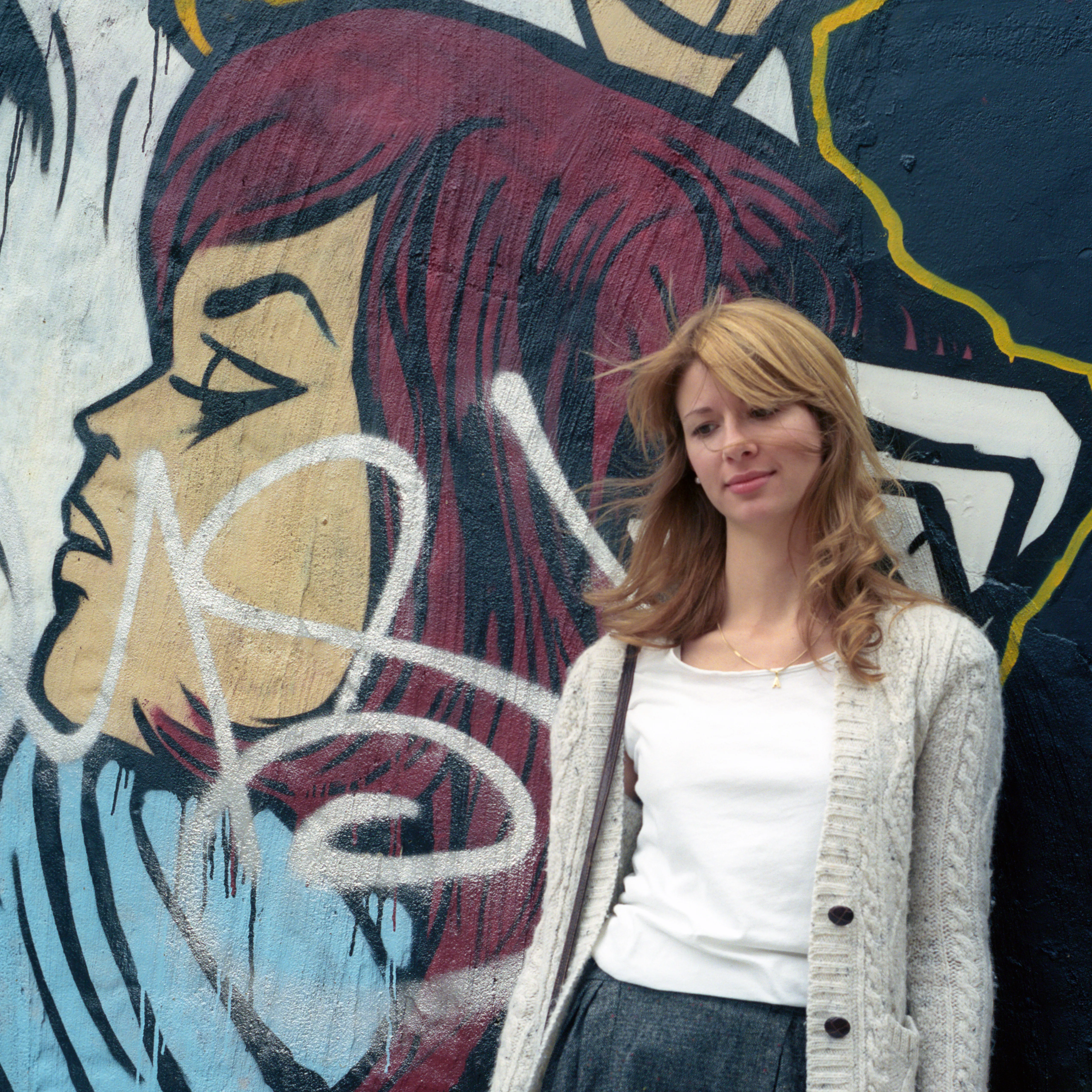 Eva standing against a painted wall mural of a woman's face, the two profiles turned away from one another.