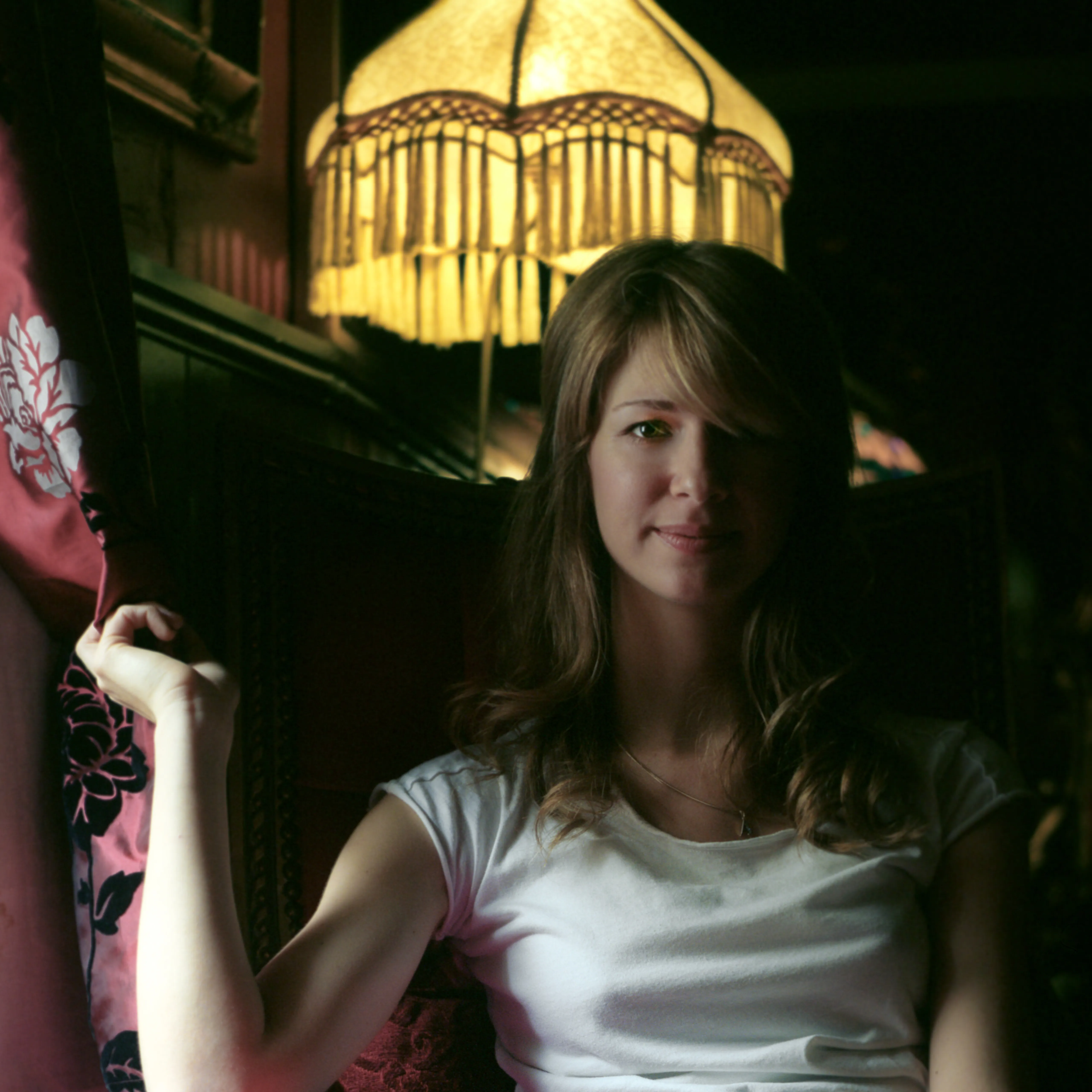 Eva in a dim interior, meeting the camera through a fall of hair, the fringed lamp lit behind her.
