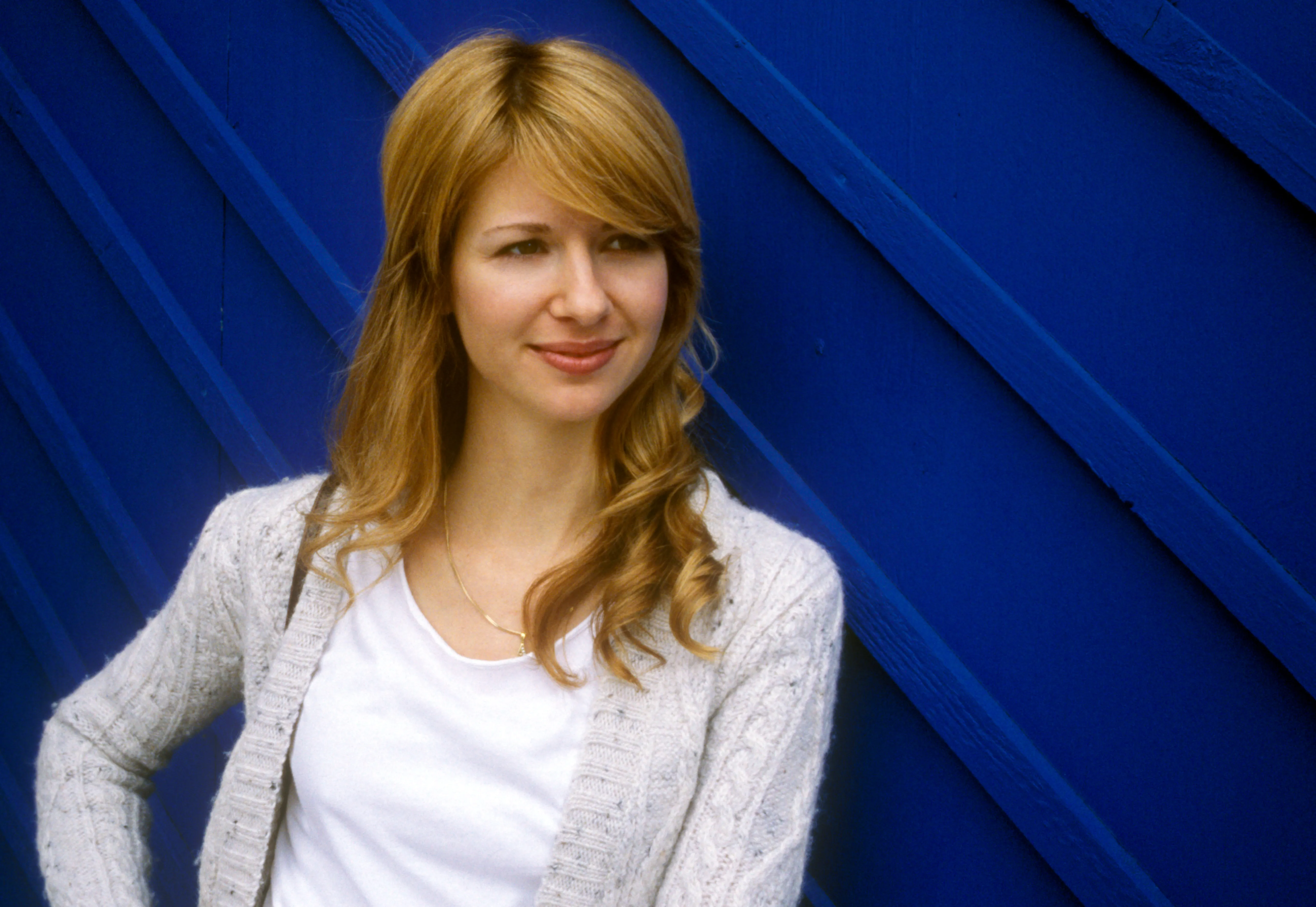 Eva against a cobalt-blue planked wall, half-turned, meeting the camera with a quiet smile.