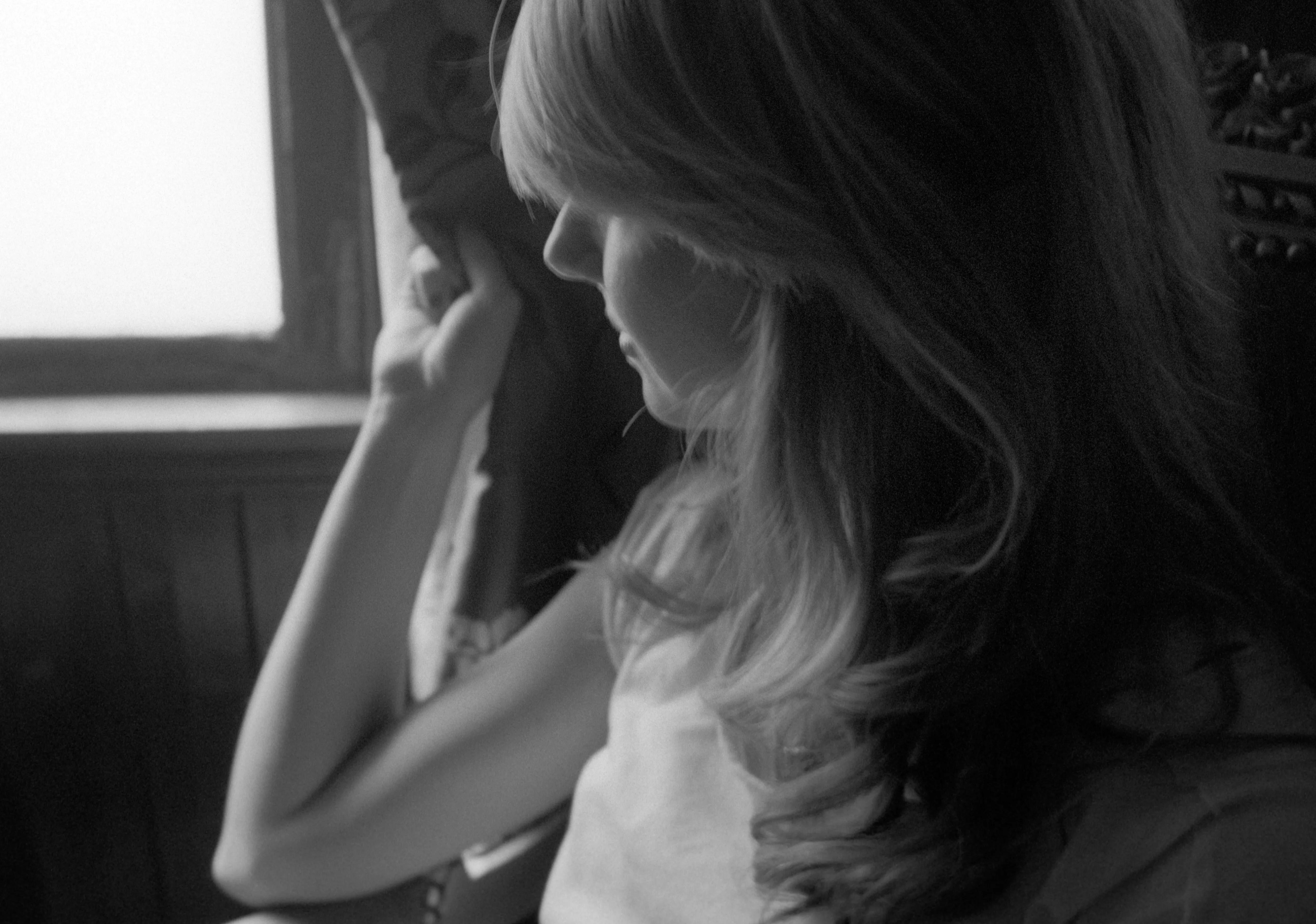 Eva in black and white, in profile by a window, head bowed with a hand raised to her face.