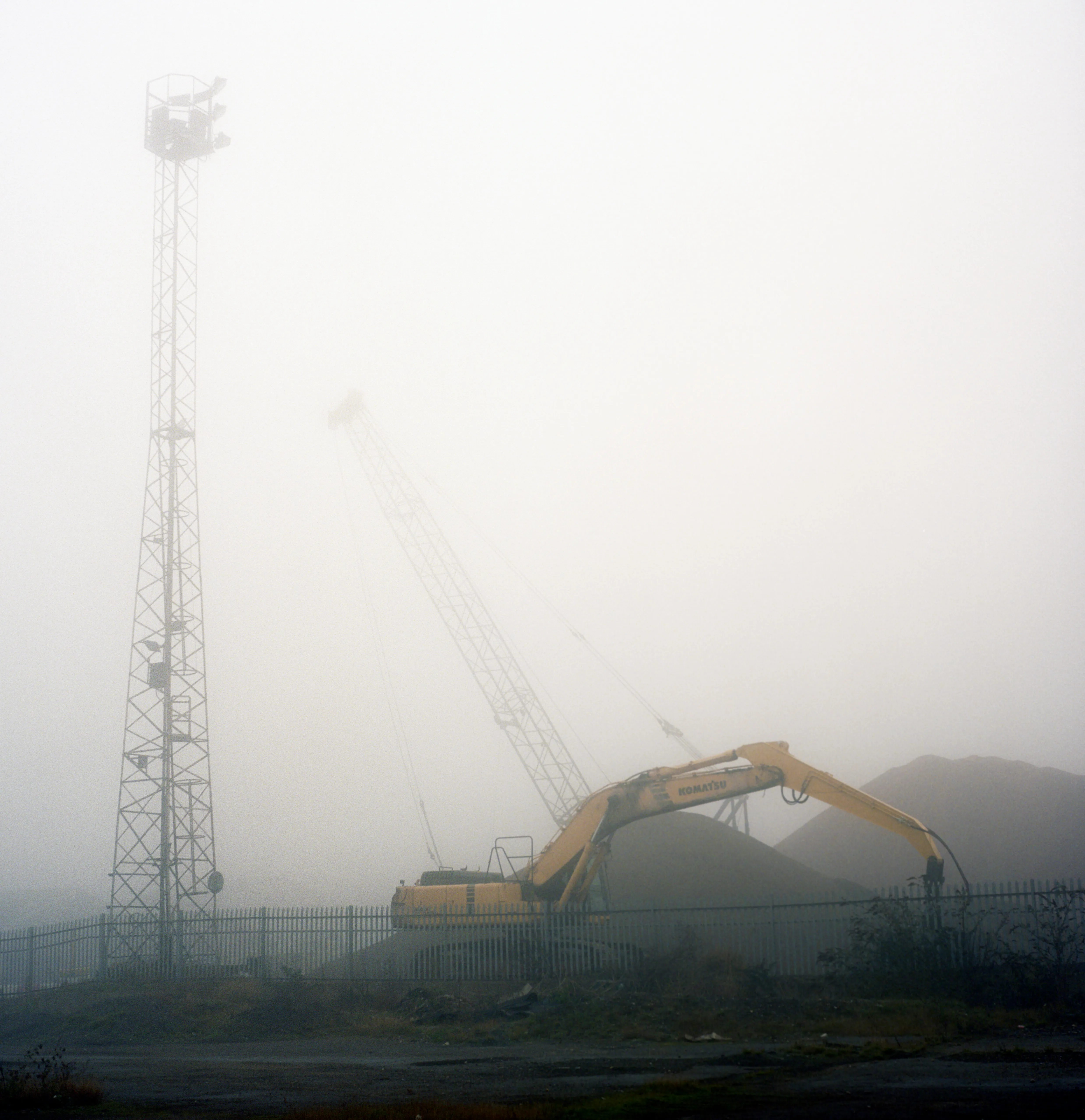 A long-reach excavator paused beside a dark spoil heap in heavy fog, a floodlight mast and a ghosted crane jib behind it.