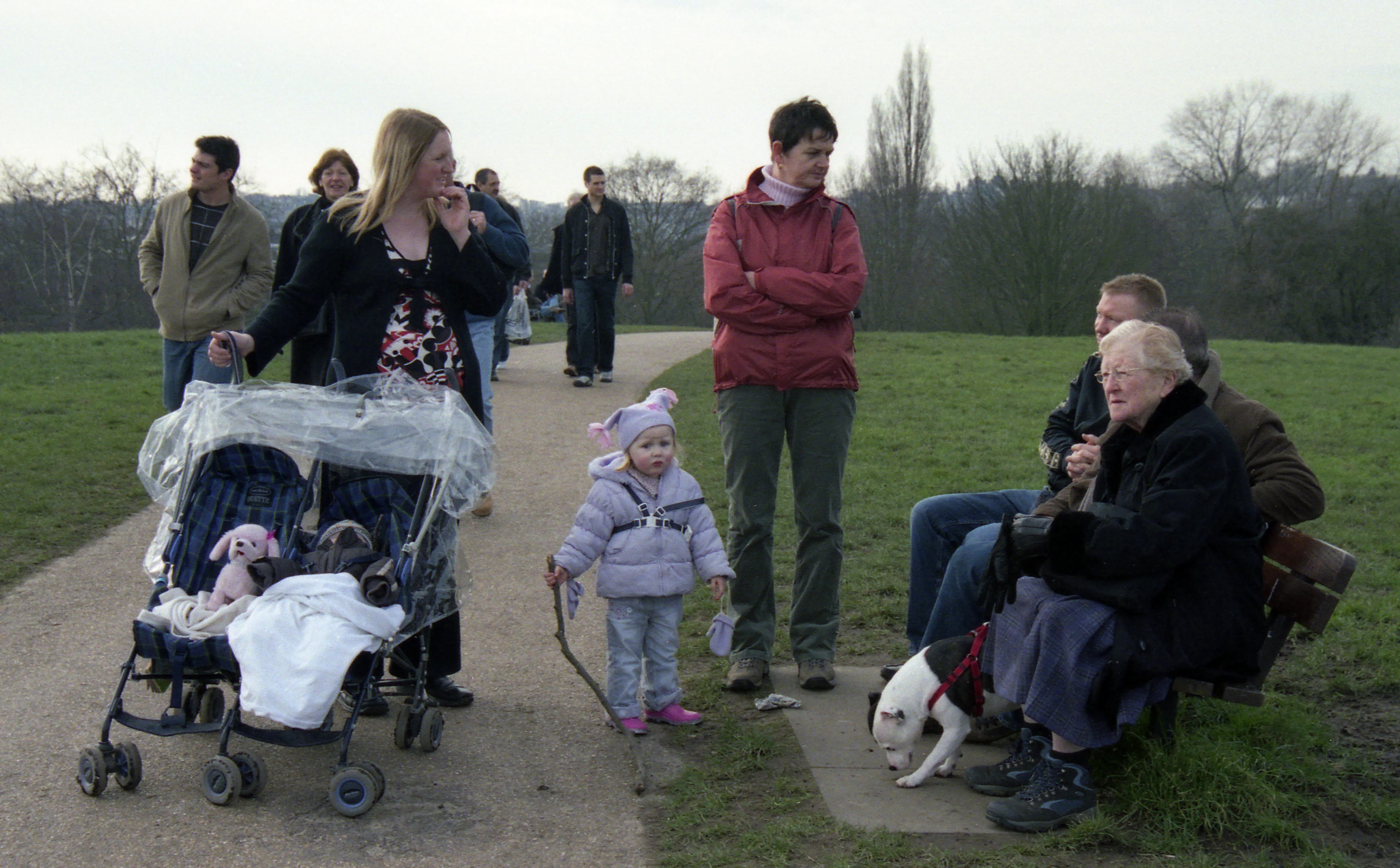 People gathered on a park path at dusk — a woman with a twin buggy, a child holding a stick, an elderly woman on a bench with a small dog.