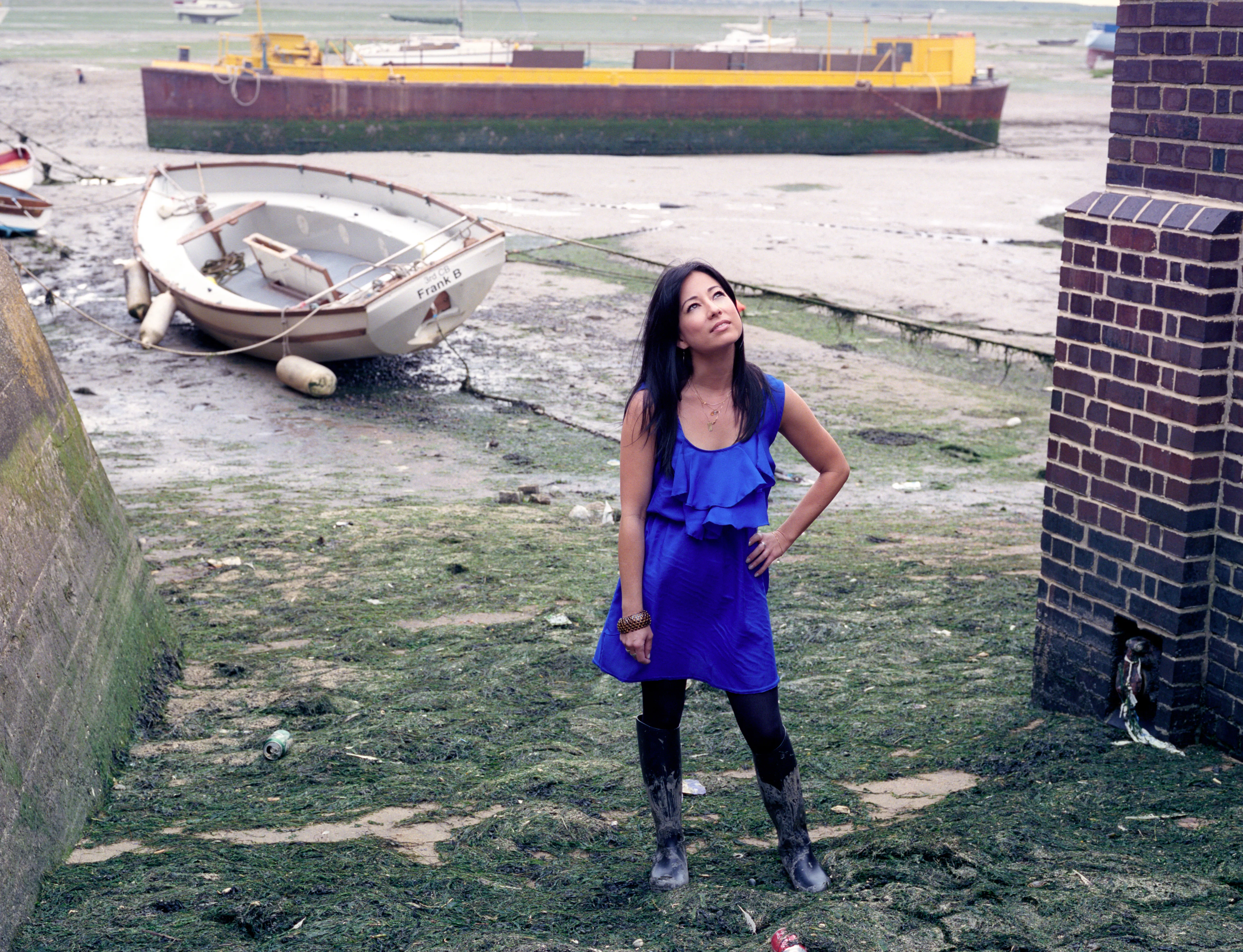 Kay in an electric-blue dress and wellington boots on an algae-covered tidal flat at low water, hand on hip, gazing upward; stranded boats and a yellow barge behind her.