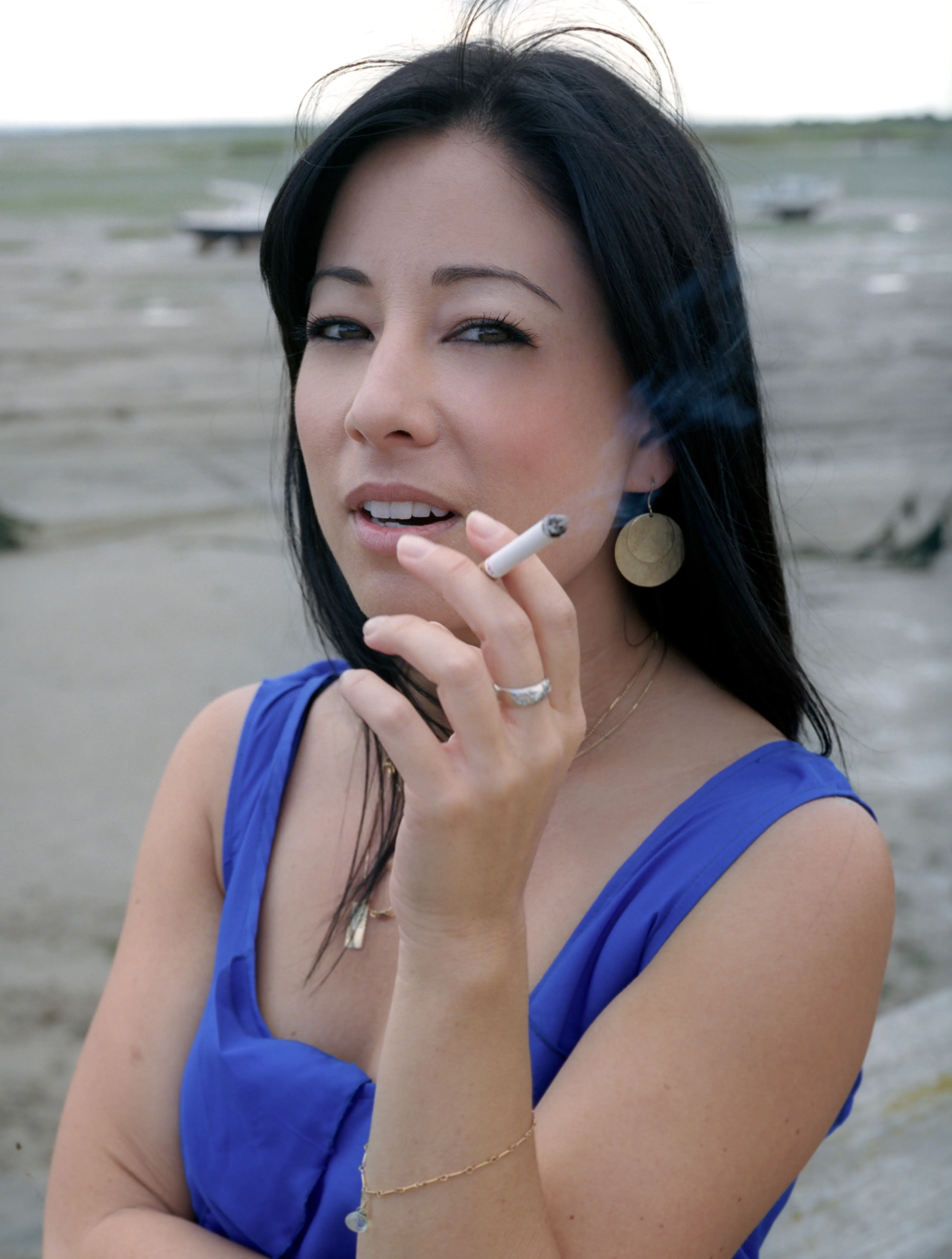 Kay in the blue dress drawing on a cigarette, smoke curling upward, glancing to the side; the pale estuary blurred behind her.