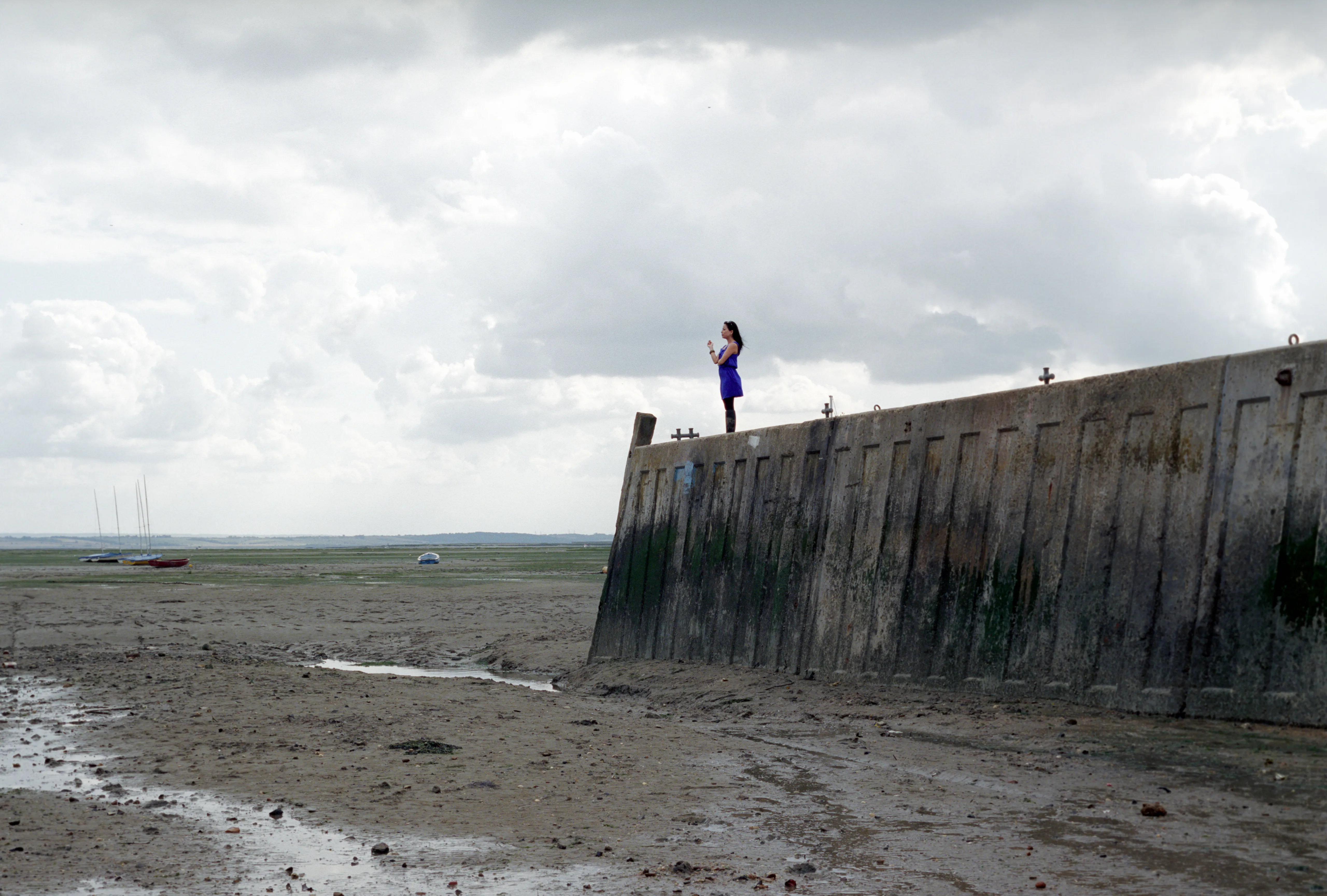 A small figure of Kay standing alone atop a long concrete sea wall, dwarfed by a vast cloud-filled sky over a drained, cracked estuary.