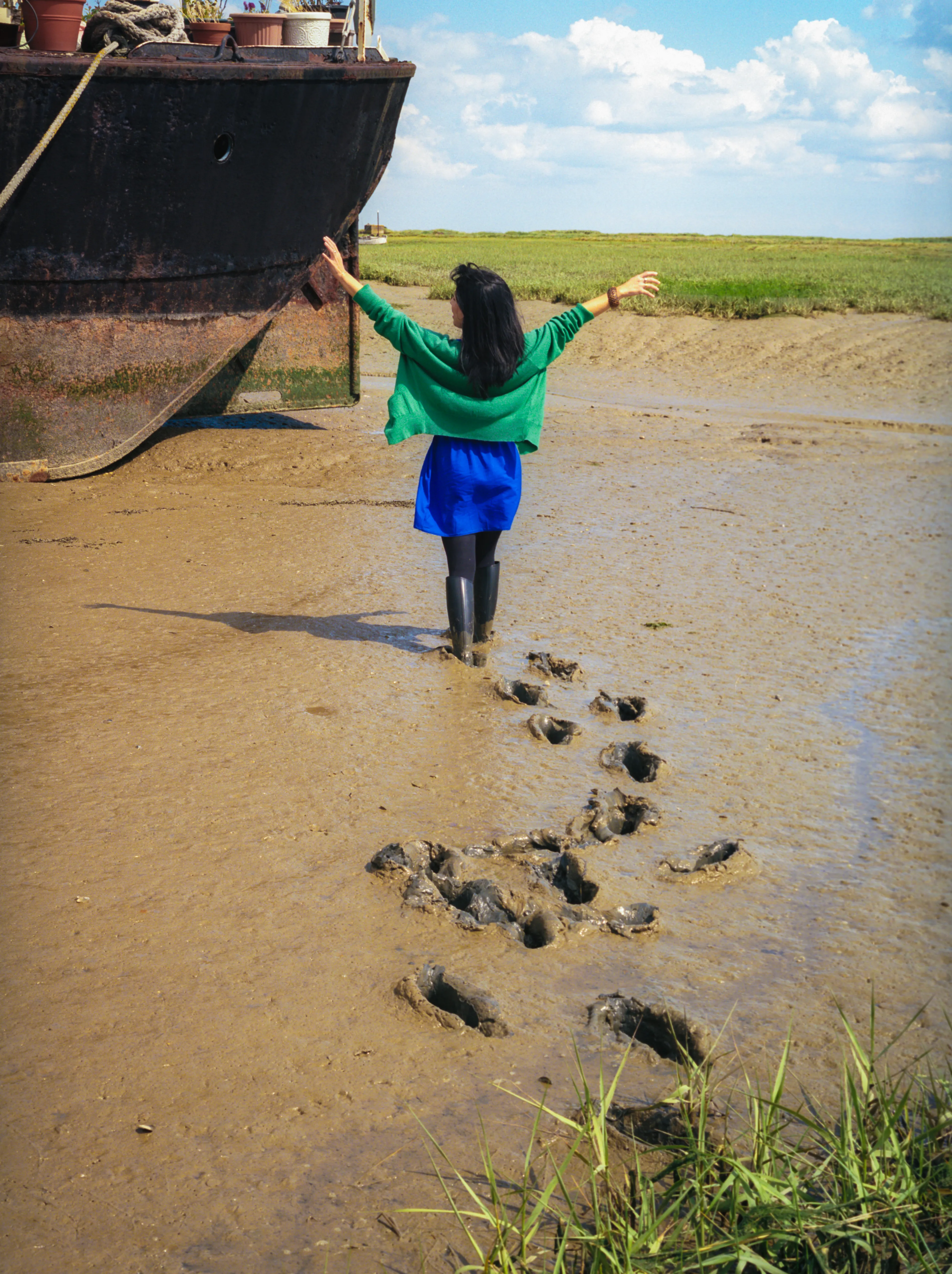 Kay seen from behind in a green cardigan and blue dress, arms outstretched, one hand on the rusted hull of a beached barge; a trail of deep footprints leads across the mud to where she stands.