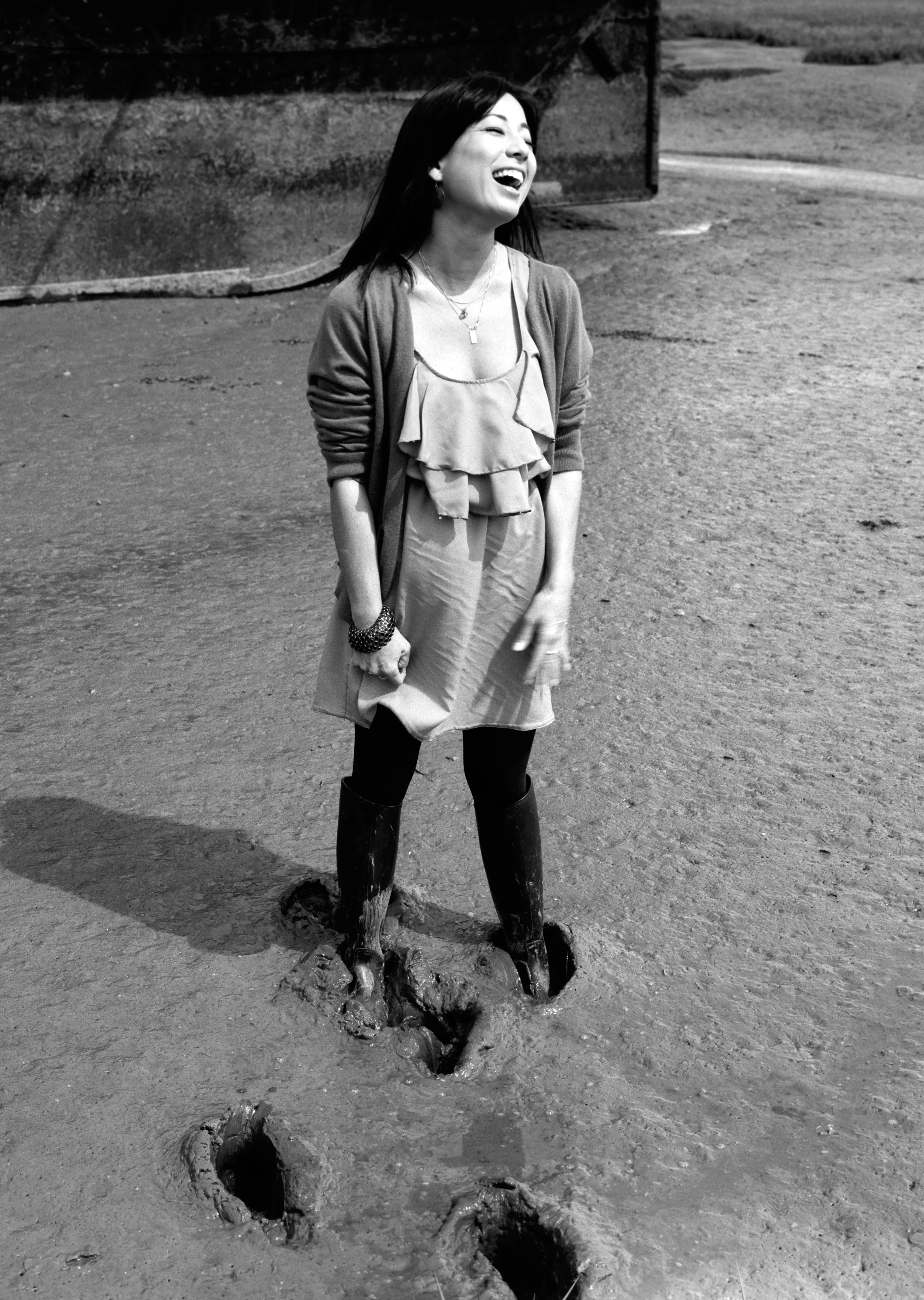 Black-and-white frame of Kay laughing with her head thrown back, boots sunk into estuary mud, footprint holes around her.