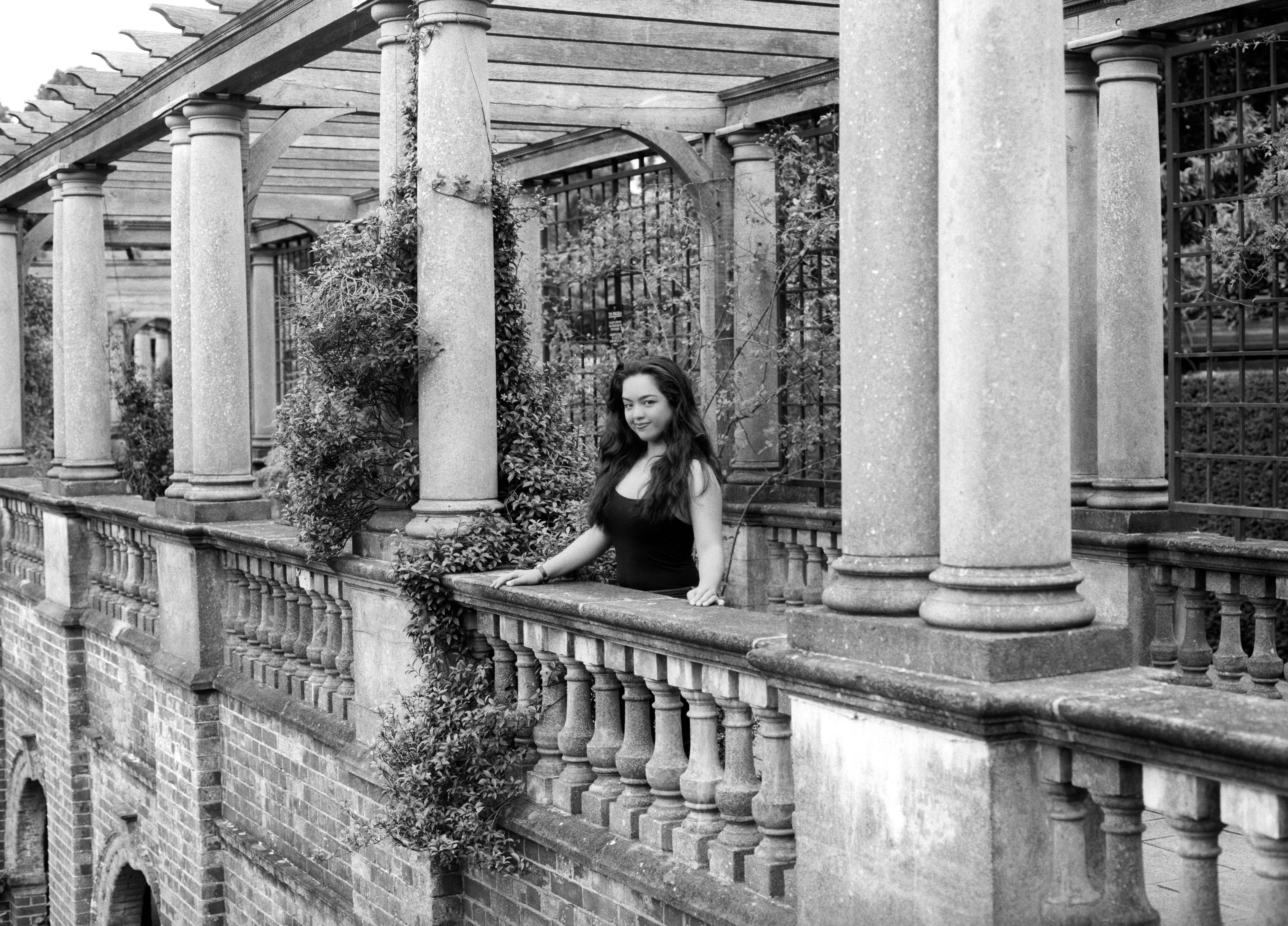 Kiana standing at a stone balustrade between the columns of a garden pergola, leaning on the rail and looking towards the camera.