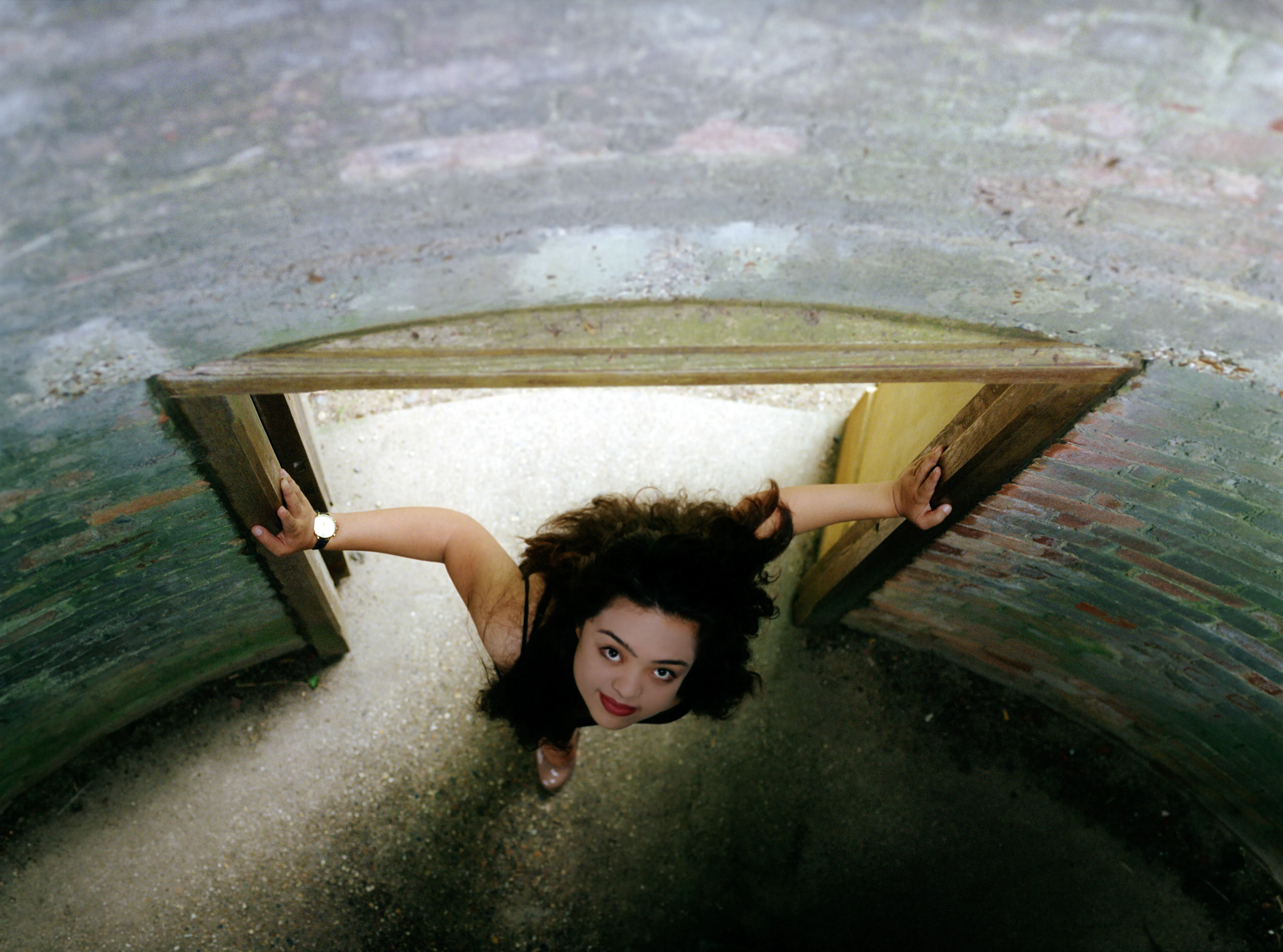 Looking down through a brick archway, Kiana stands in the opening below with both arms braced against the timber door frames, her face turned up to the camera.