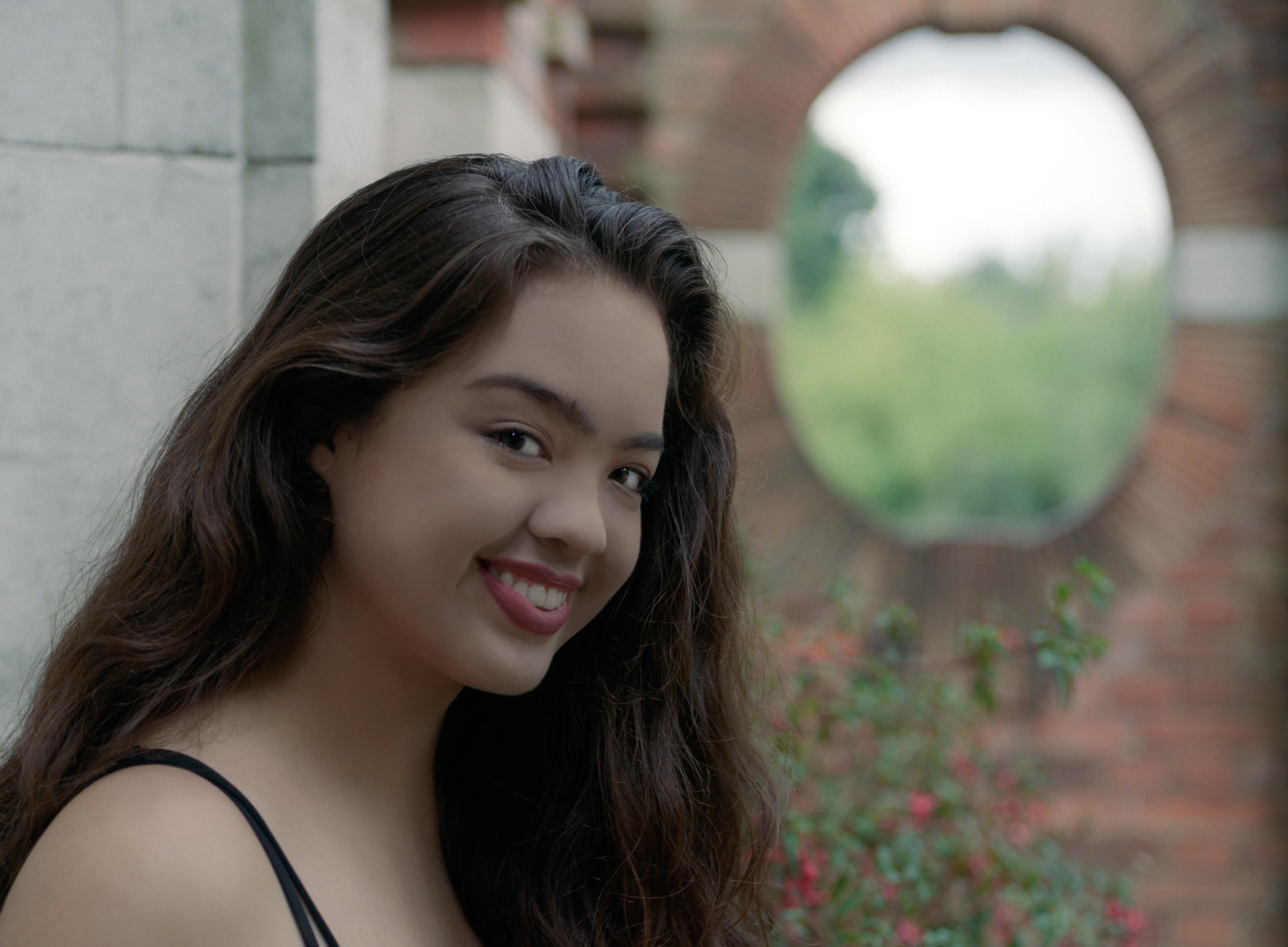 Kiana in three-quarter view beside a stone wall, long hair over one shoulder, a circular brick window and greenery behind her.