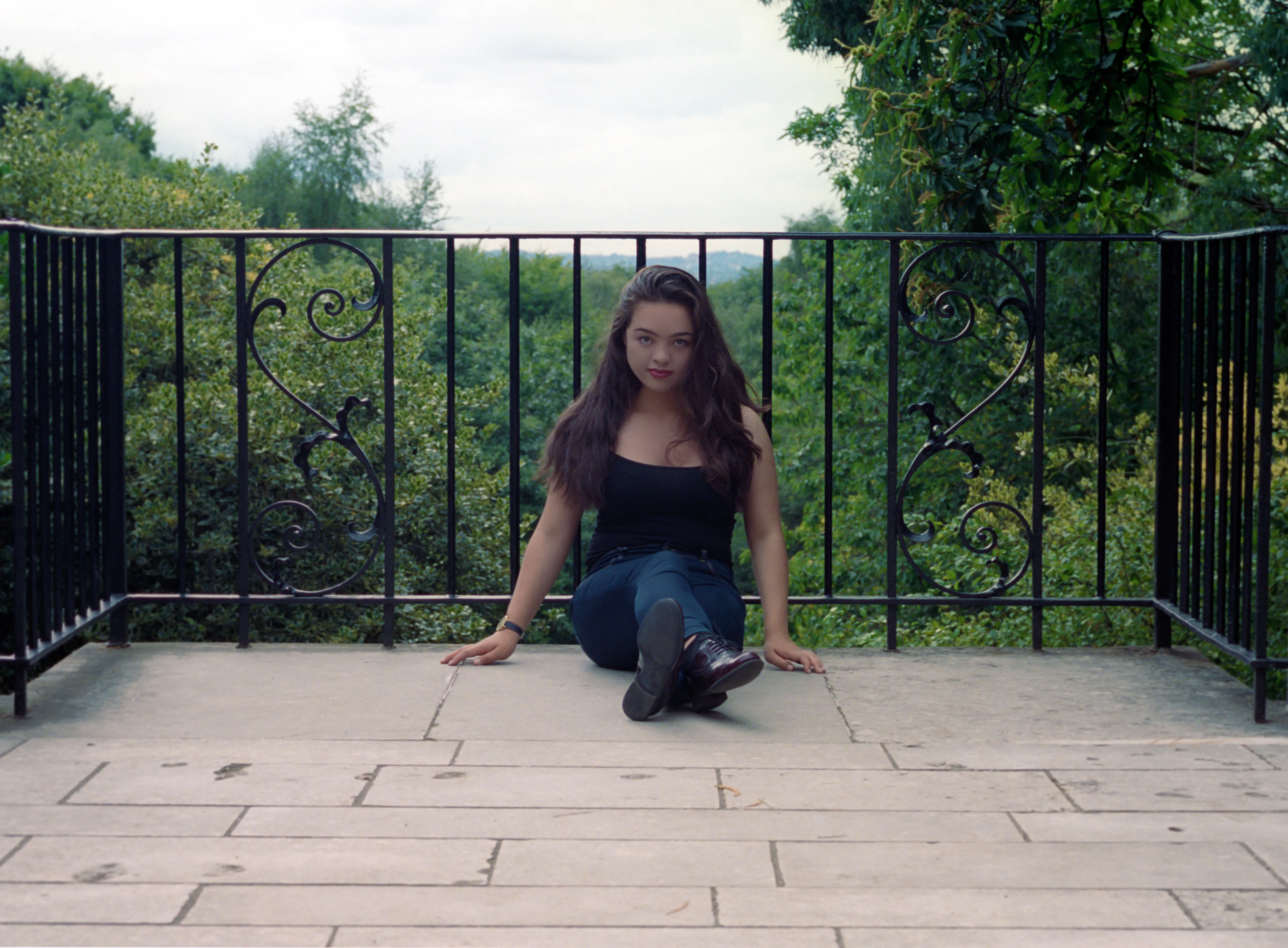Kiana seated on stone flagstones before a wrought-iron railing with scrolled panels, hands on the ground, facing the camera.