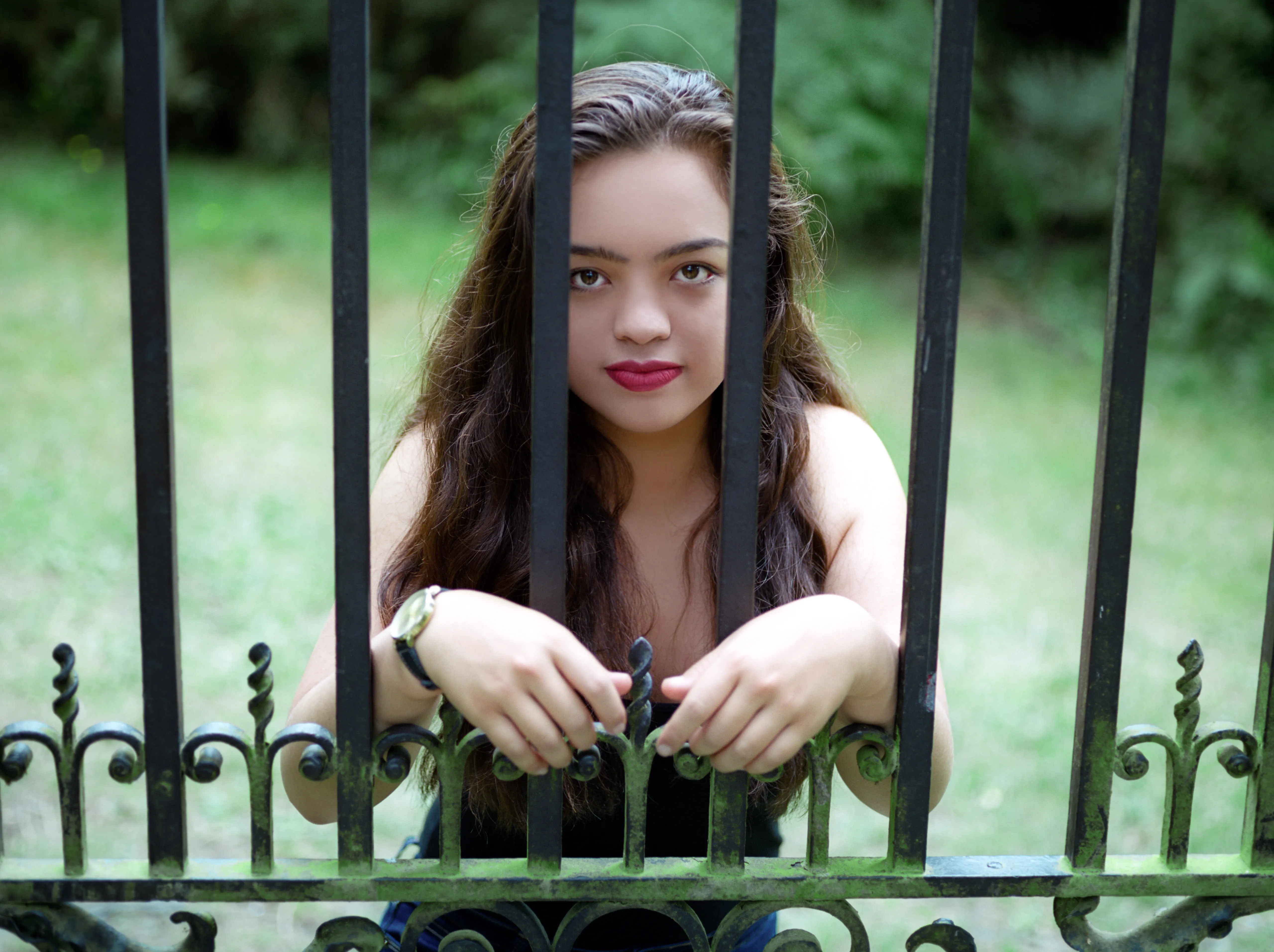 Kiana behind a tall wrought-iron gate, hands resting on the bars, looking through them towards the camera.