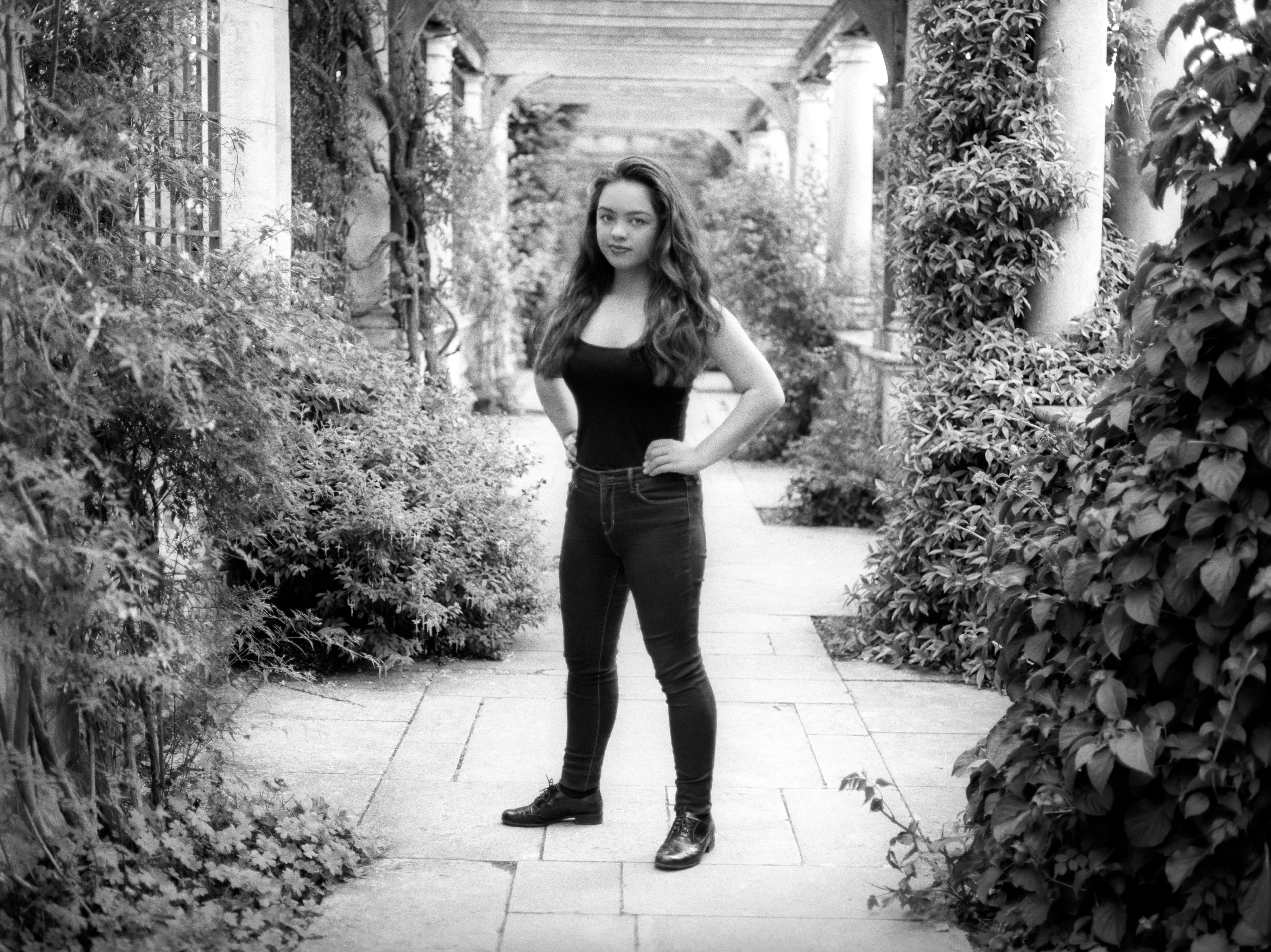 Kiana standing on the paved path of a garden pergola, hands on her hips, columns and foliage receding behind her.