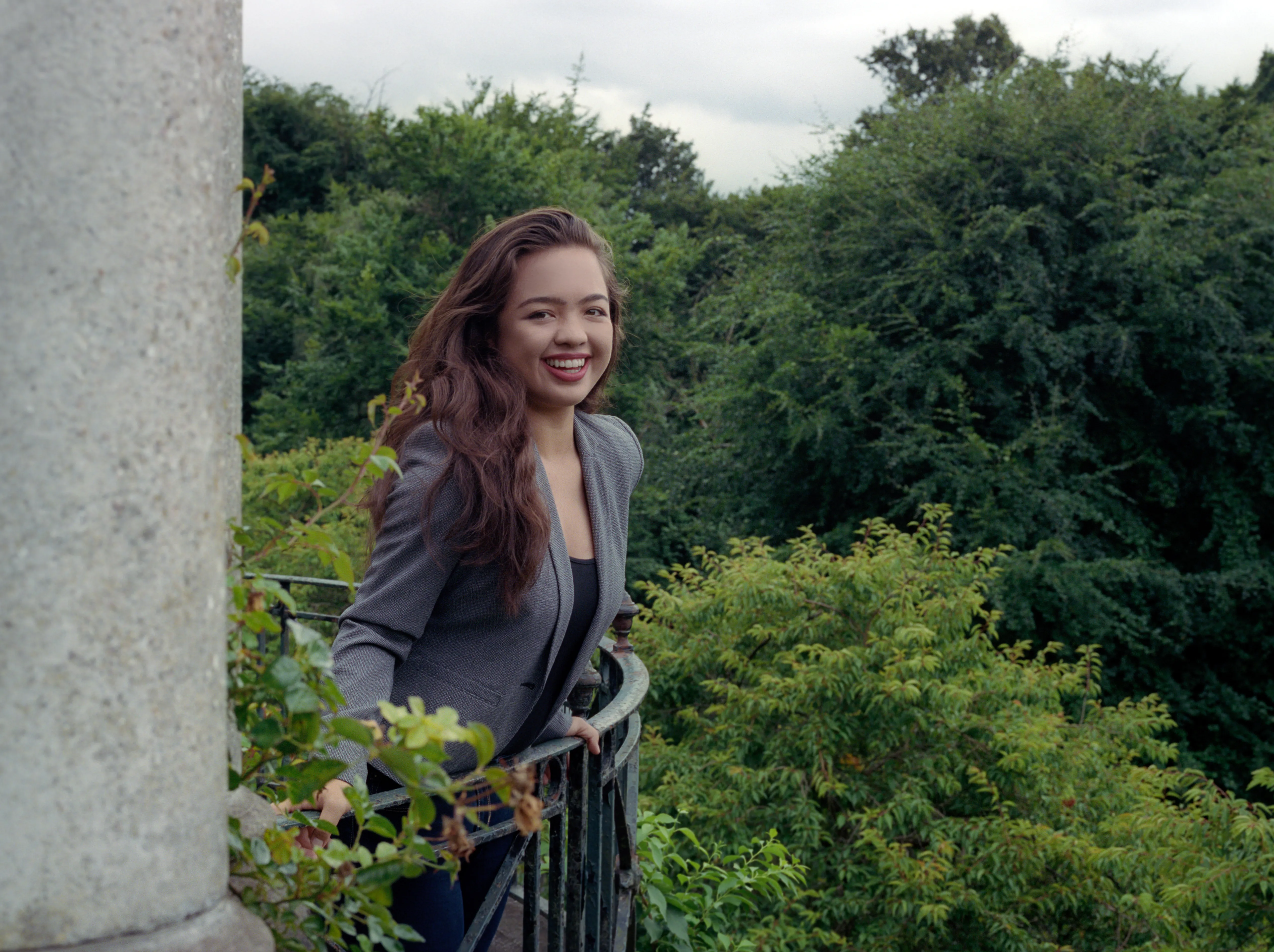 Kiana standing at a railing at the edge of a wooded terrace, wearing a grey blazer, a stone column beside her on the left, smiling.