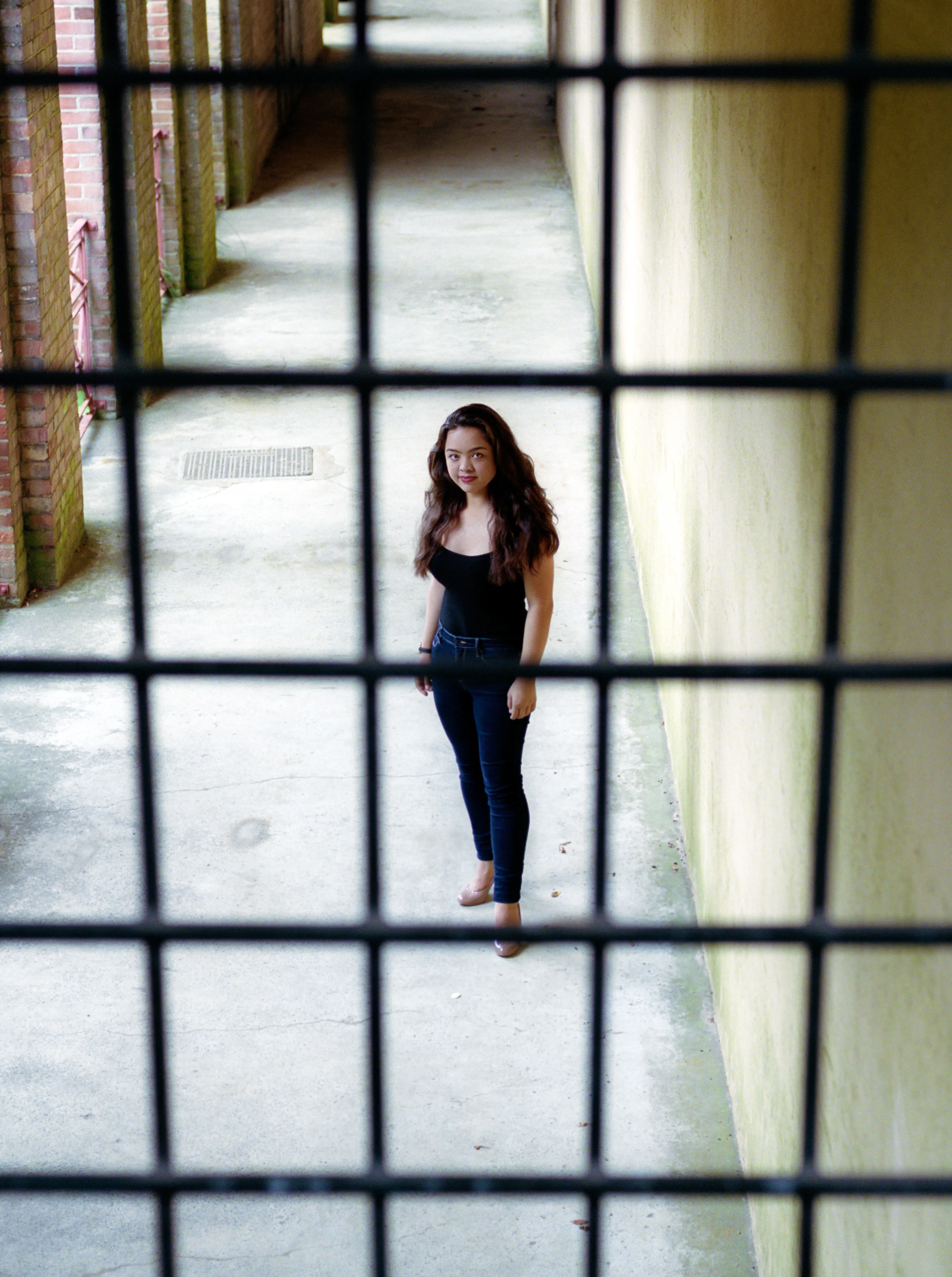 Seen from above through a metal grille, Kiana stands in a paved passage between brick and rendered walls, looking up towards the camera.