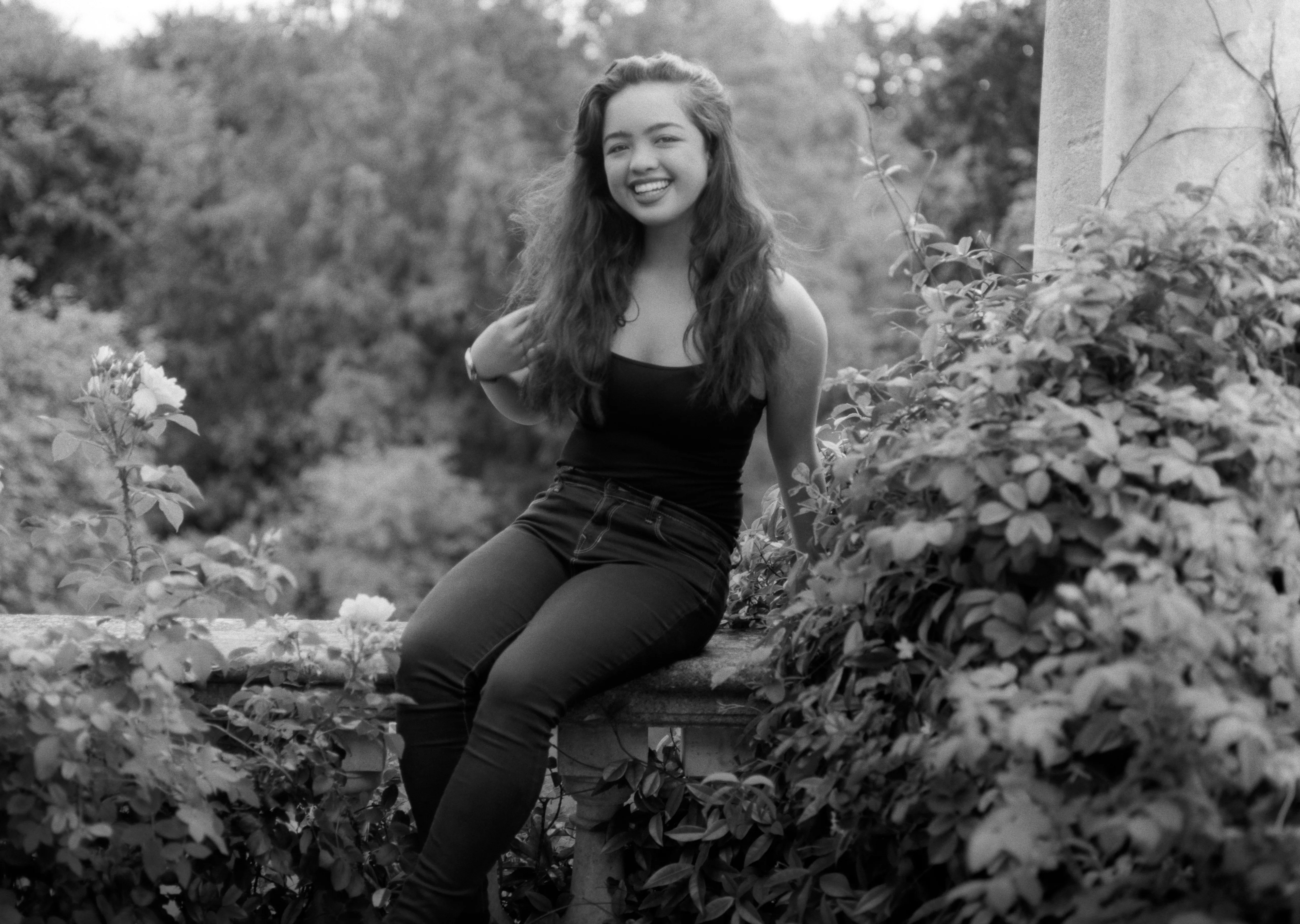 Kiana seated on a stone balustrade in a garden, one hand raised to her hair, rose bushes beside her and a pillar to the right, laughing.