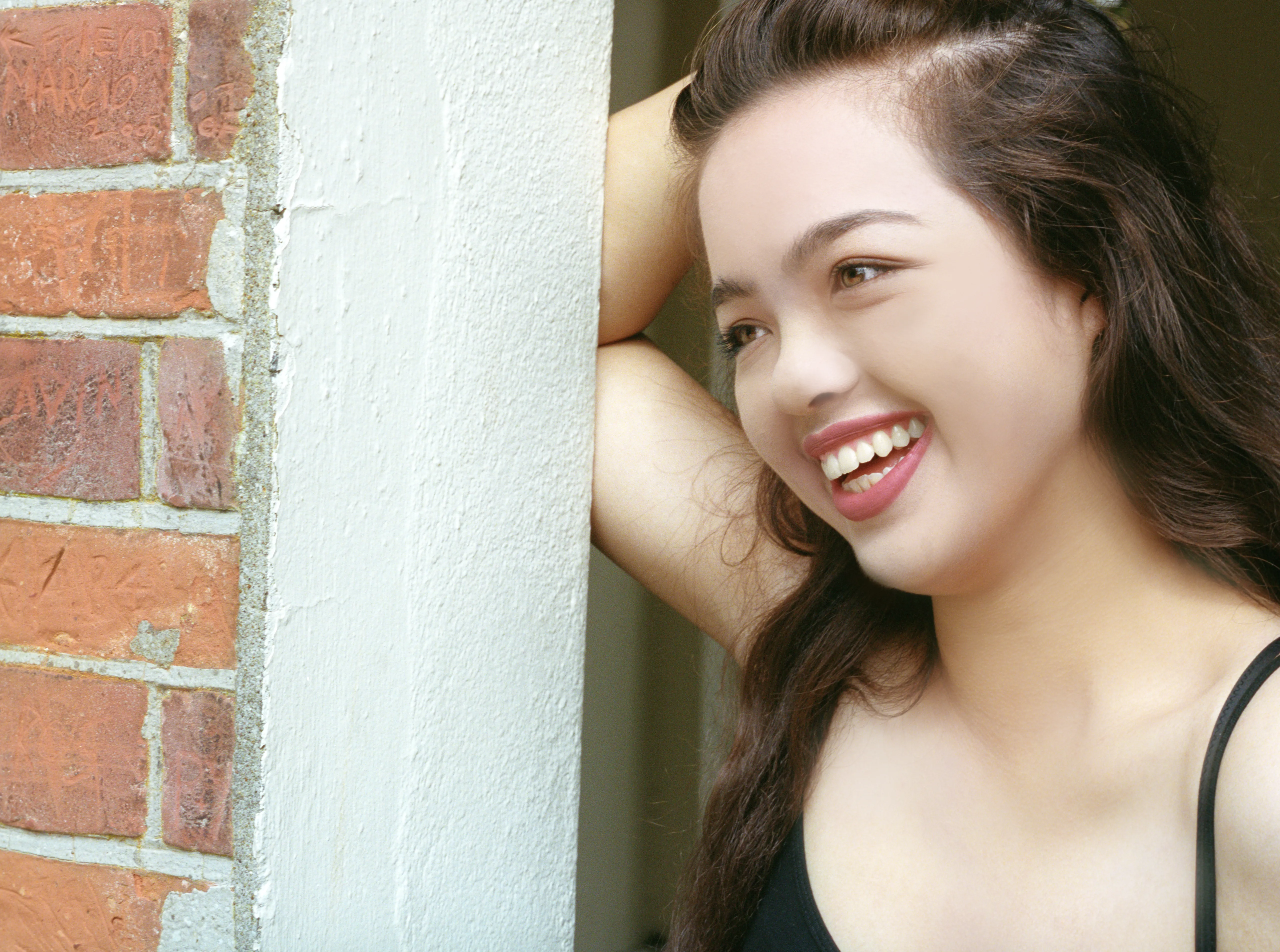 Kiana leaning against a painted brick corner, one arm raised behind her head, laughing and looking to the side.