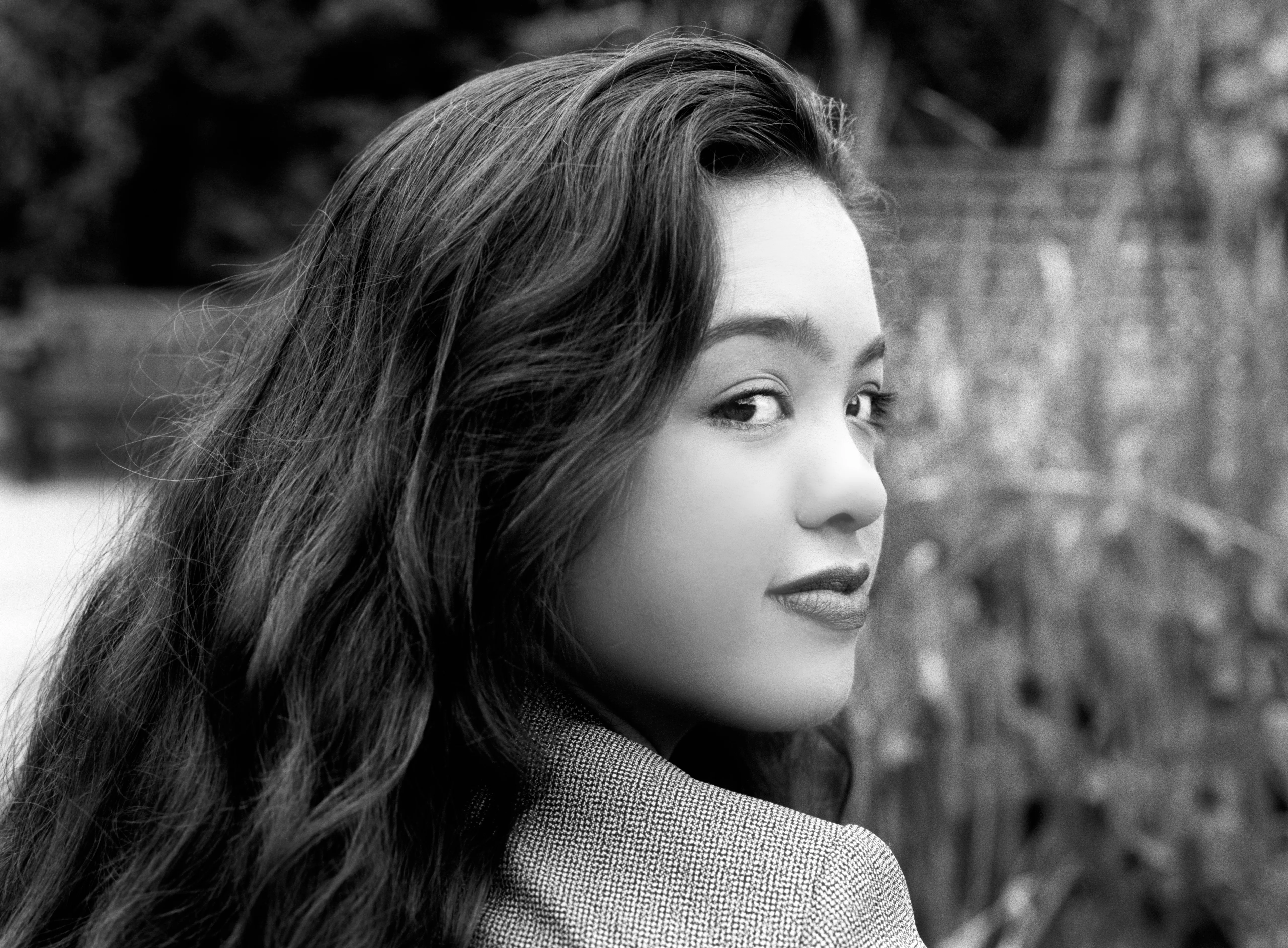 Kiana glancing back over her shoulder towards the camera, long hair falling down her back, wearing a herringbone jacket.