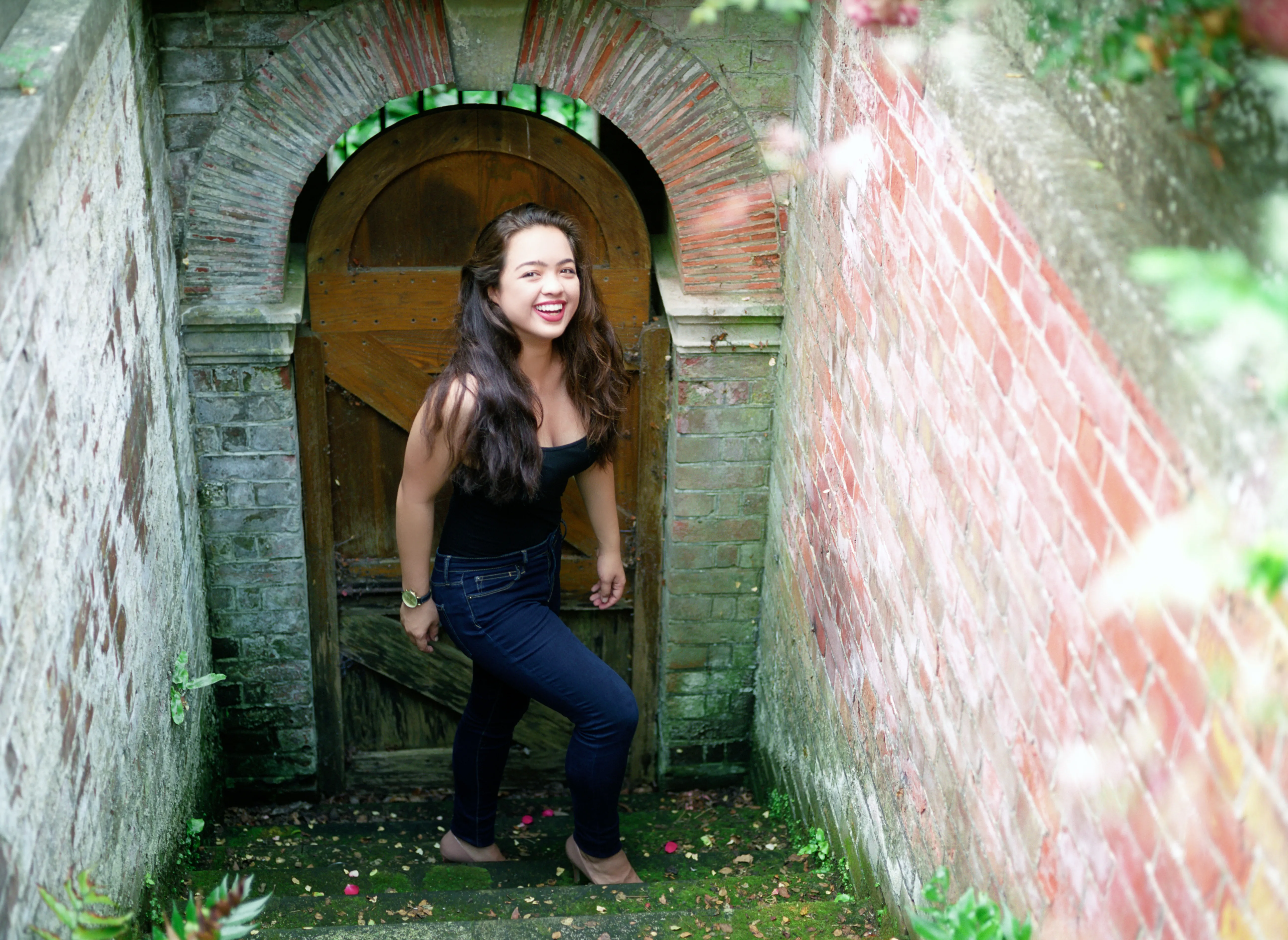 Kiana at an arched timber door at the foot of a narrow brick stairway, the brick walls converging towards her, laughing.