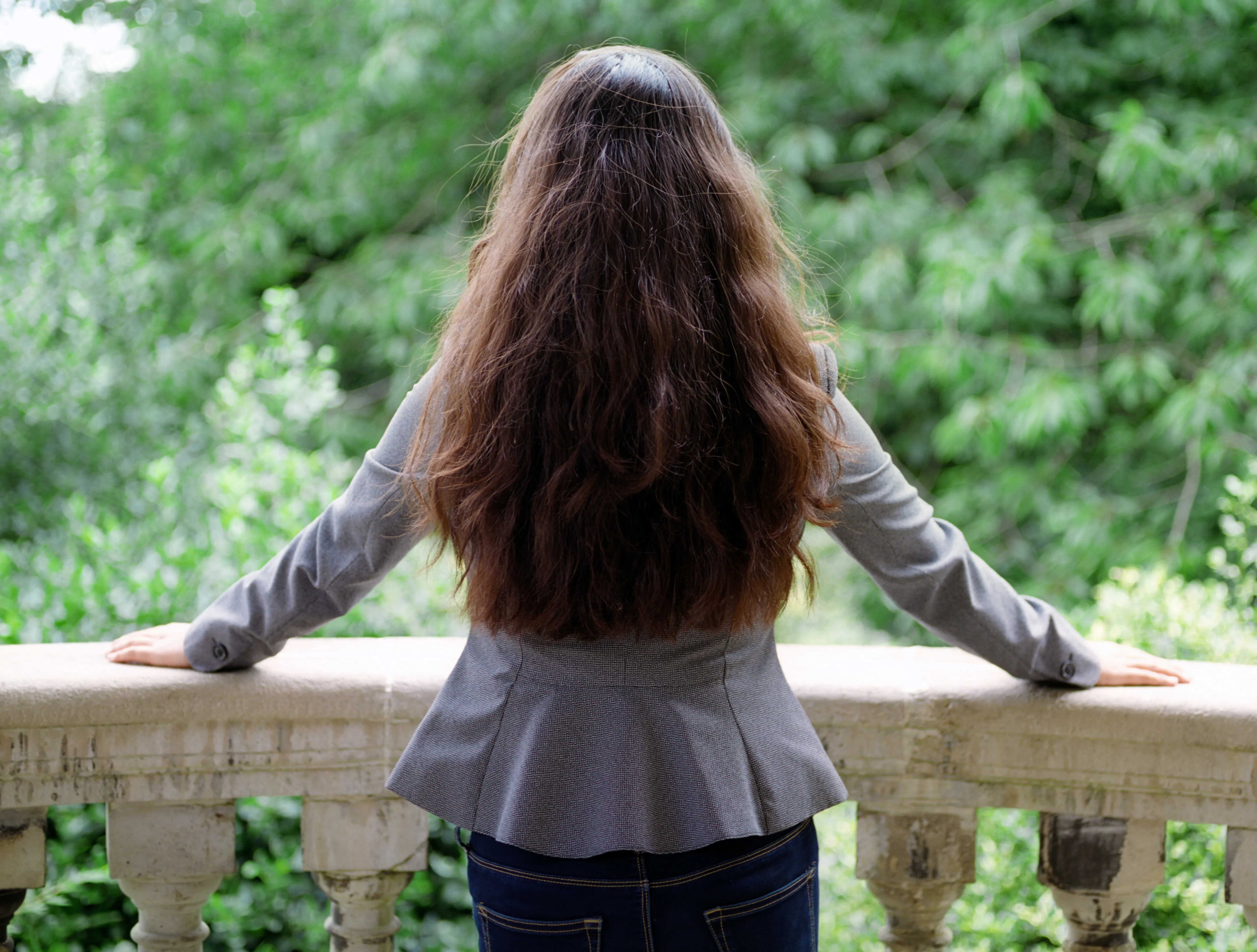 Kiana seen from behind at a stone balustrade, arms spread wide to grip the rail, facing a wall of green foliage.