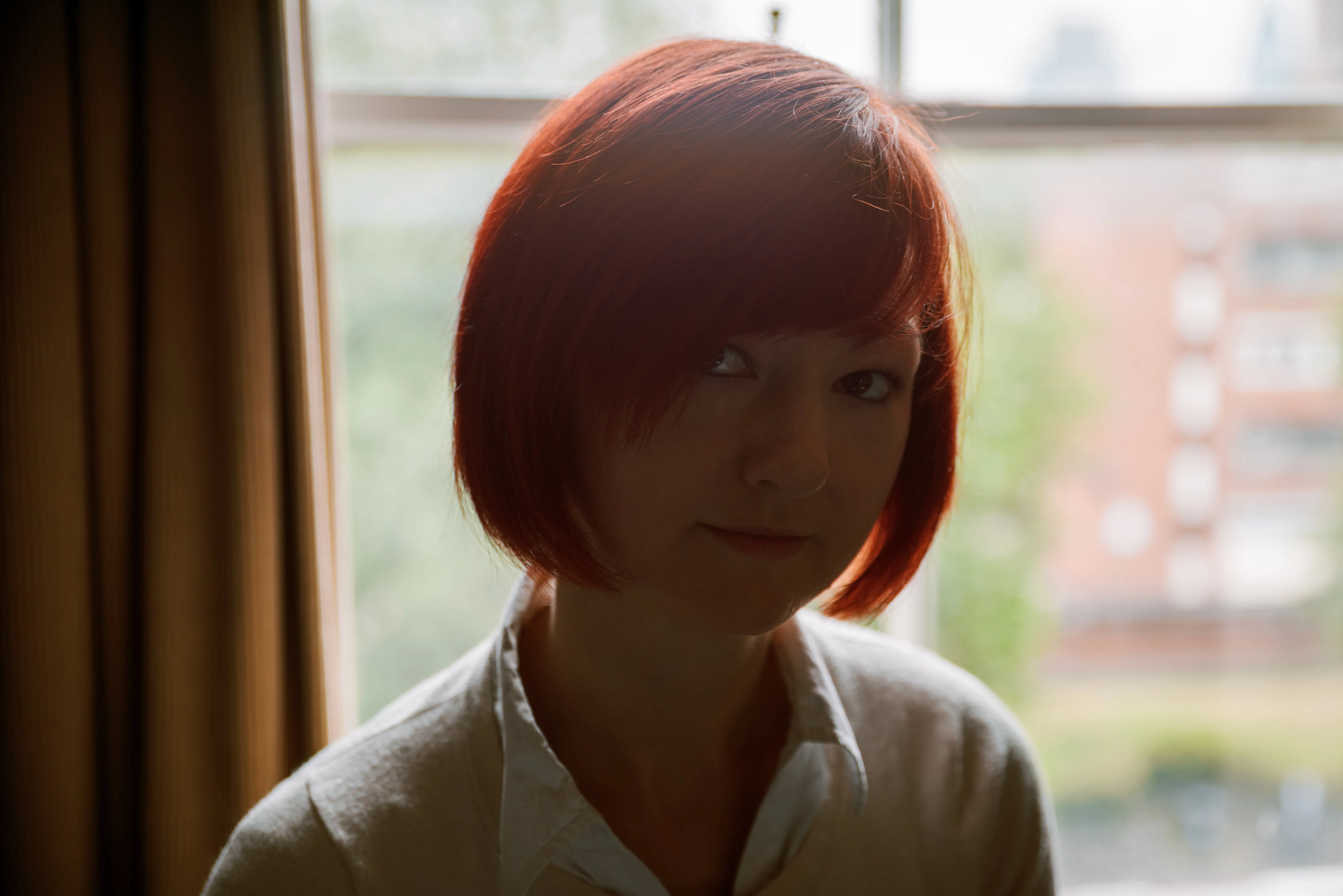 A woman with an auburn bob in a white shirt, backlit by a window, looking down and away.