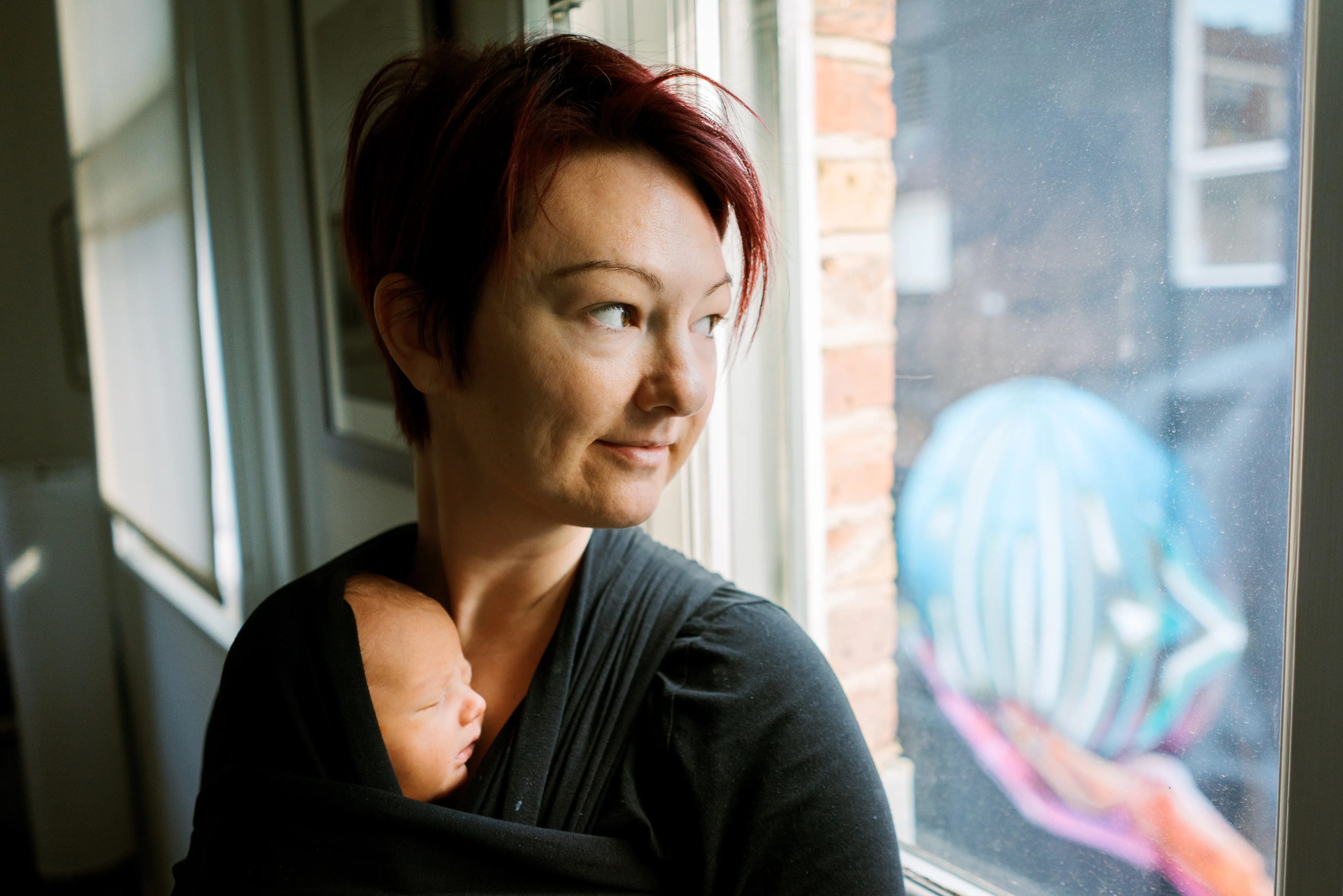 A woman with red hair holds a sleeping newborn against her chest in a wrap, looking out of a window in soft daylight.