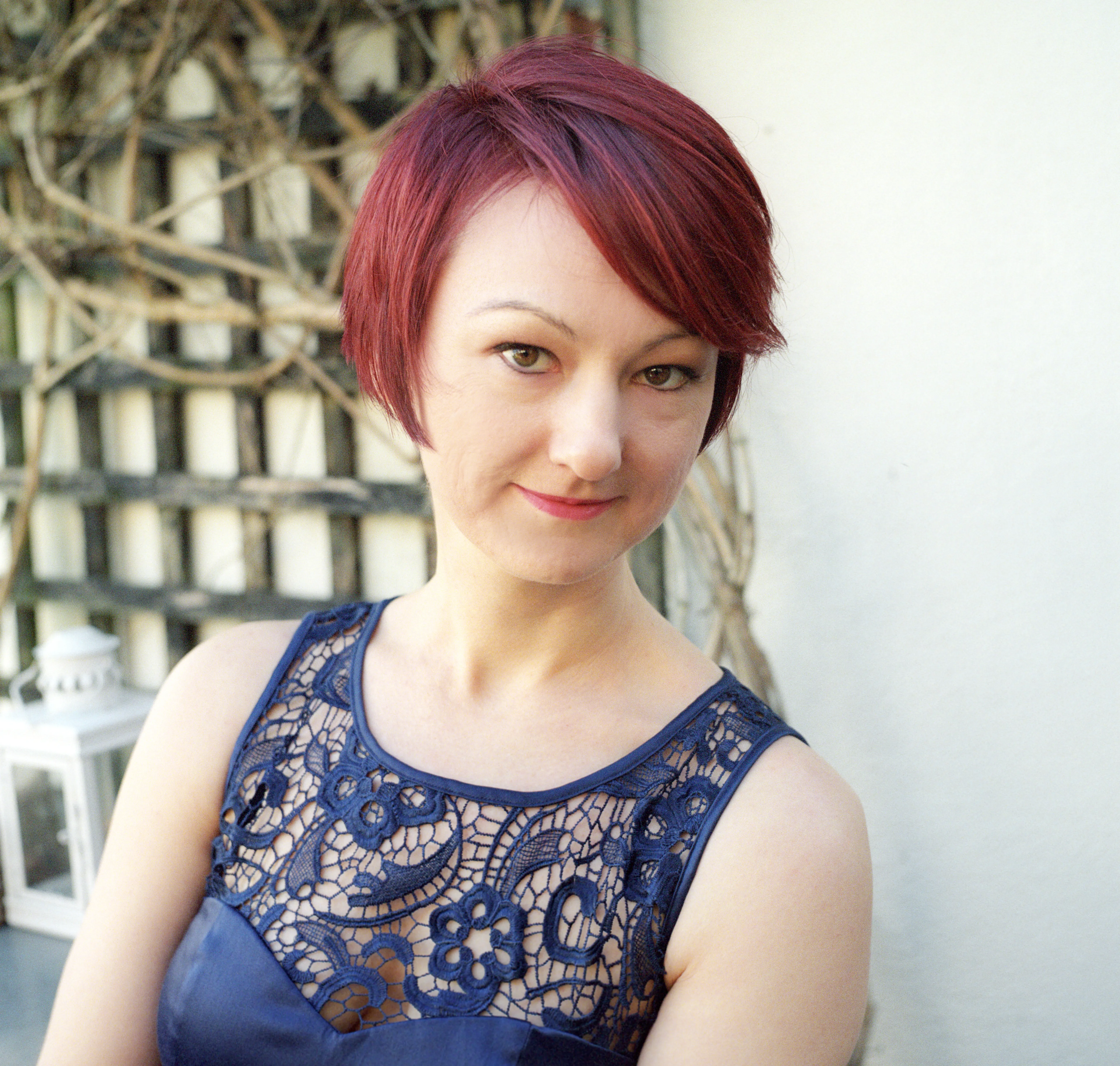 A woman with red hair in a navy lace dress, seated outdoors against a white wall and bare trellis, smiling at the camera.