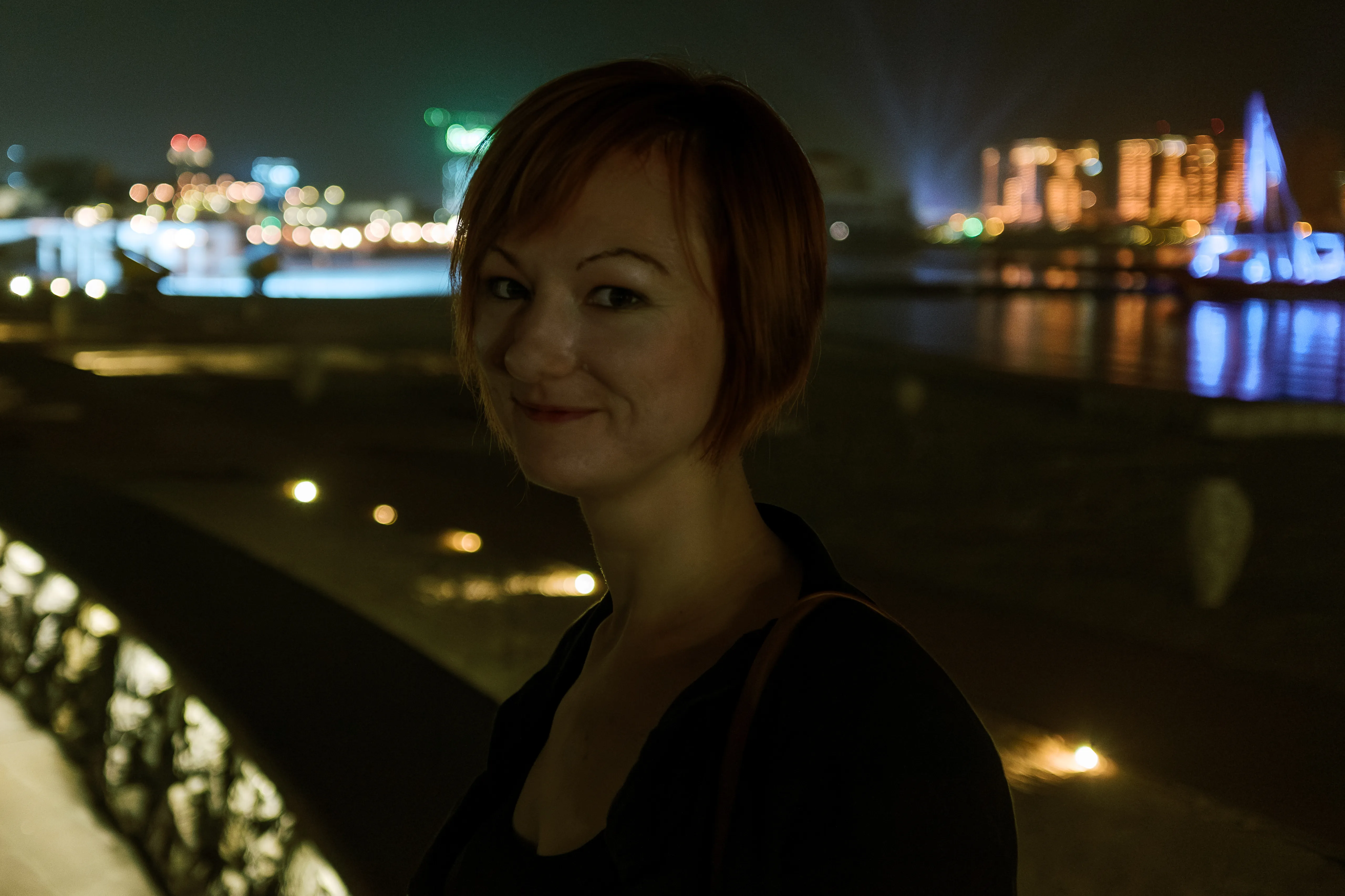 A woman in dark clothing at night, turned to the camera, with out-of-focus city lights across water behind her.