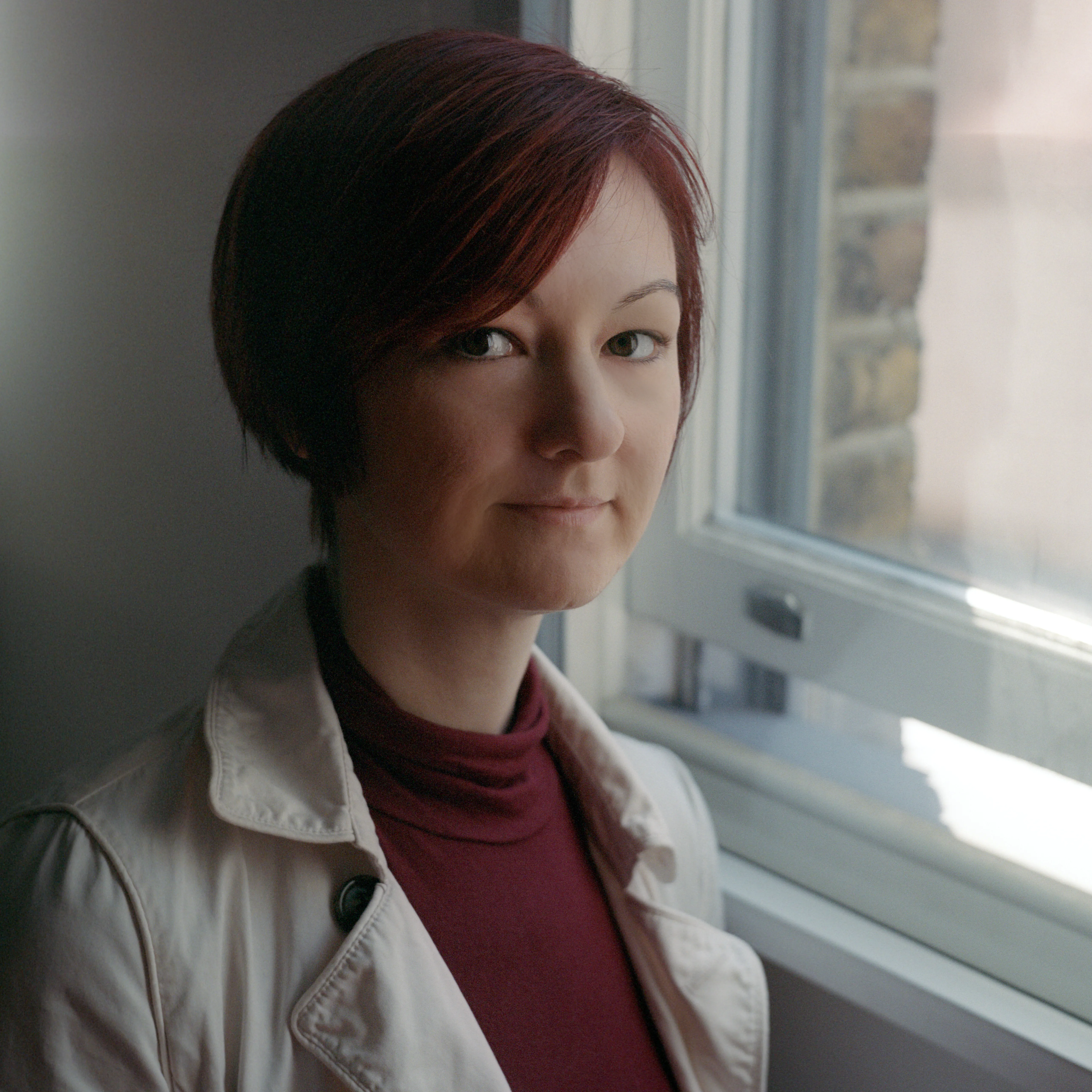 A woman with dark red hair in a pale coat and burgundy roll-neck, seated by a window, looking towards the camera.