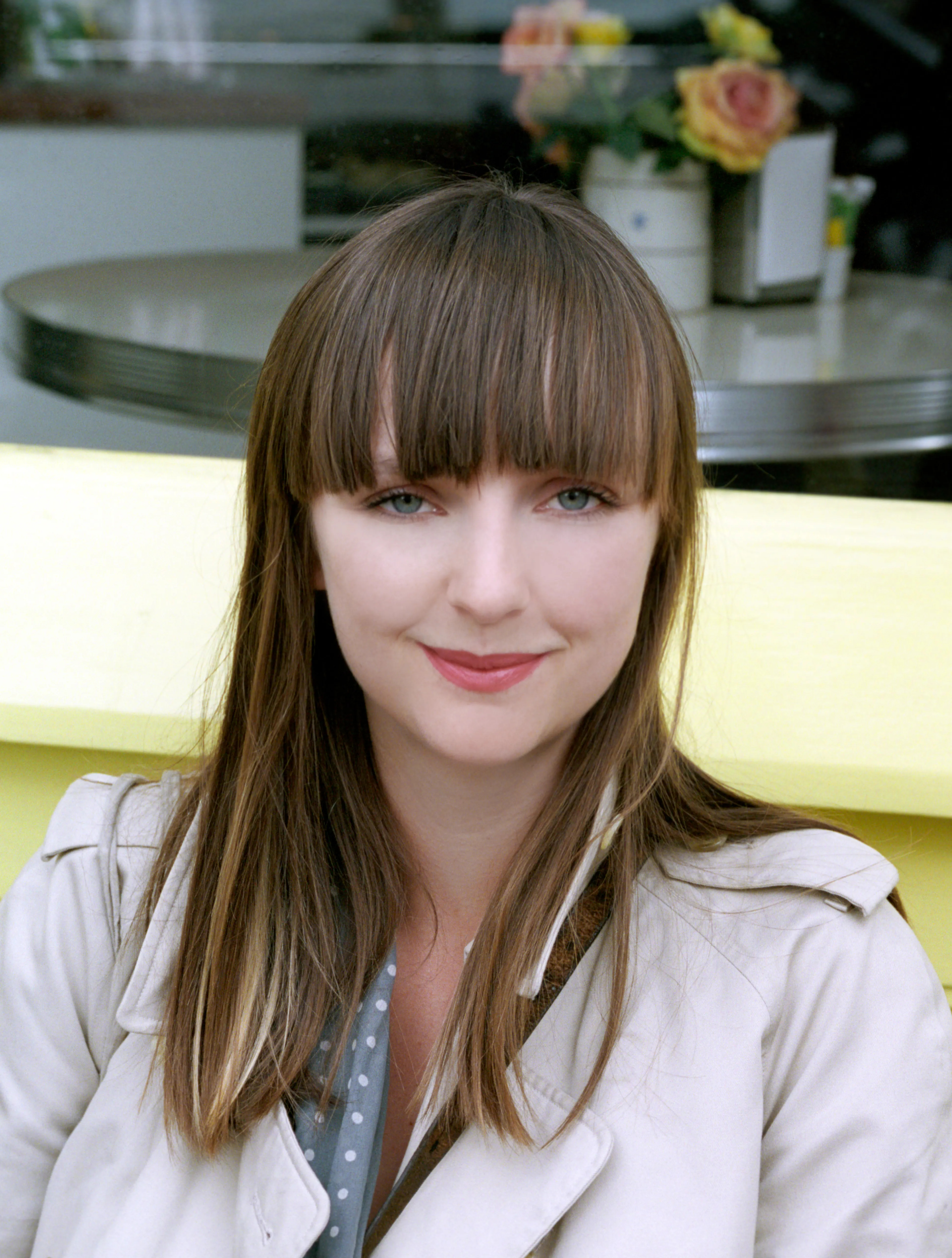 Lauren in a beige trench coat over a grey polka-dot top, facing the camera with a slight smile; a cafe table with roses behind her.