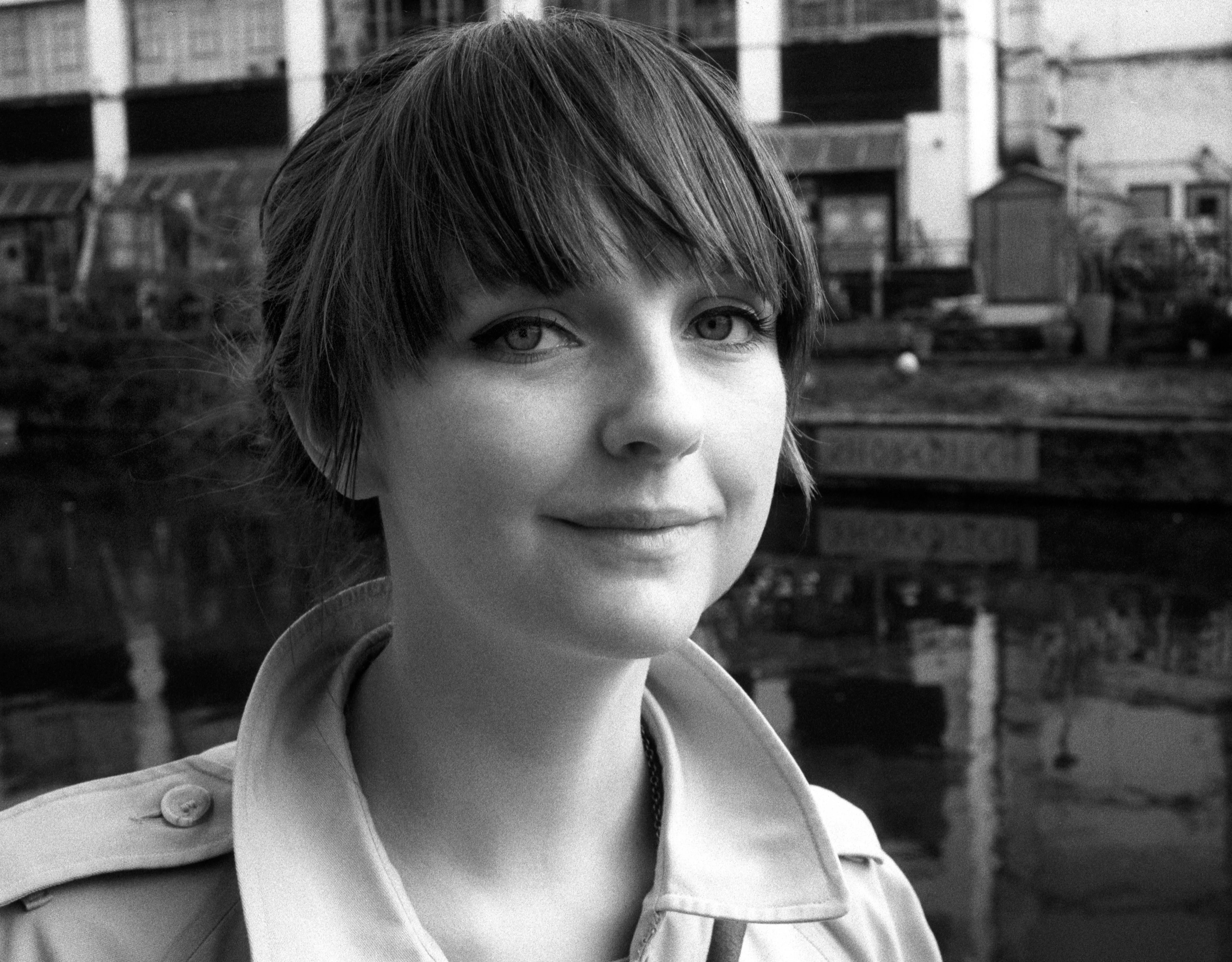 Lauren, hair up with a dark fringe, looking directly at the camera with a faint smile beside a canal, dark trees and water behind her. Black and white.
