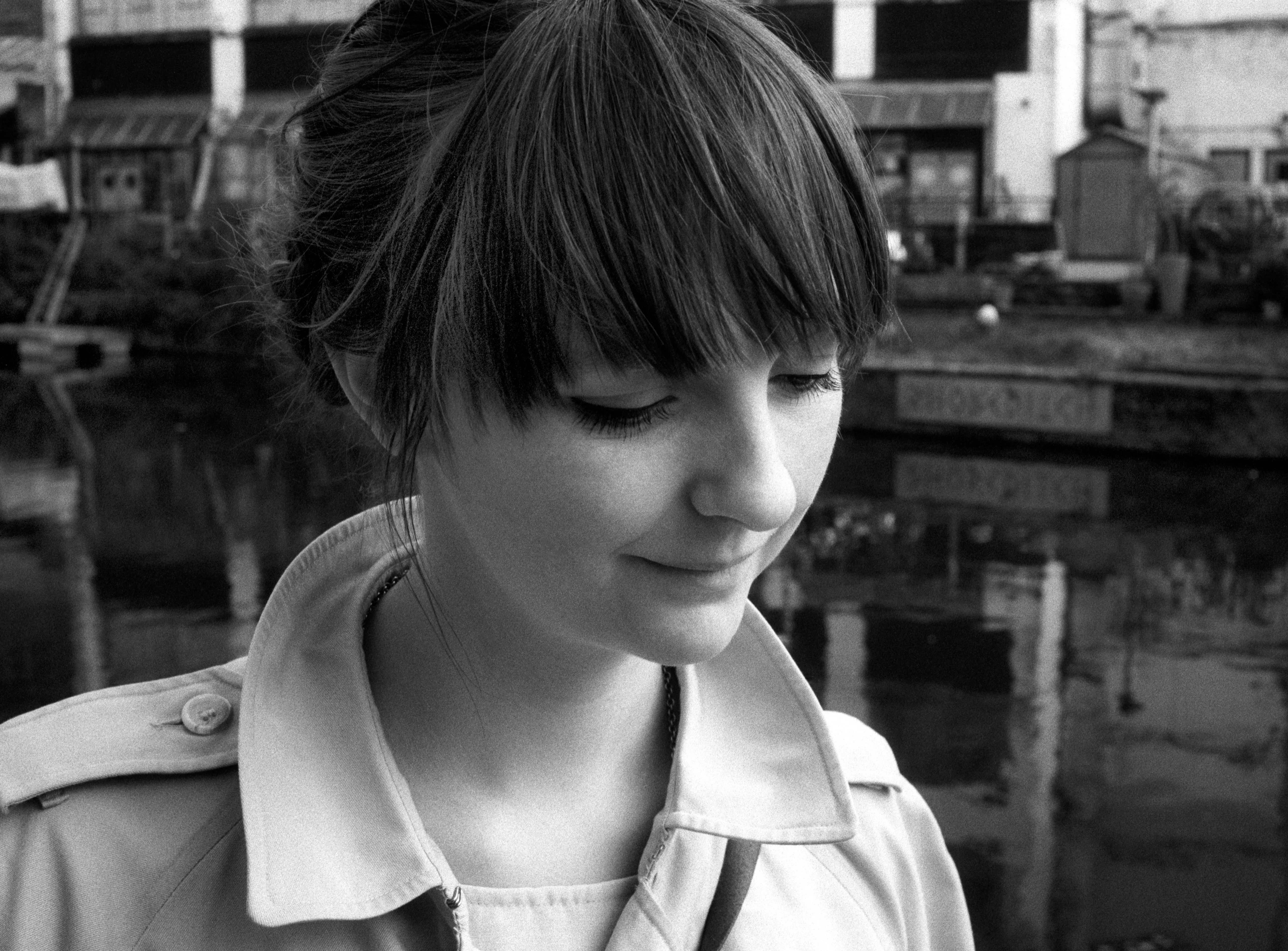 Lauren, hair up with a dark fringe, in a pale trench coat, looking down with a faint smile beside a canal, with industrial buildings reflected in the still water behind her. Black and white.