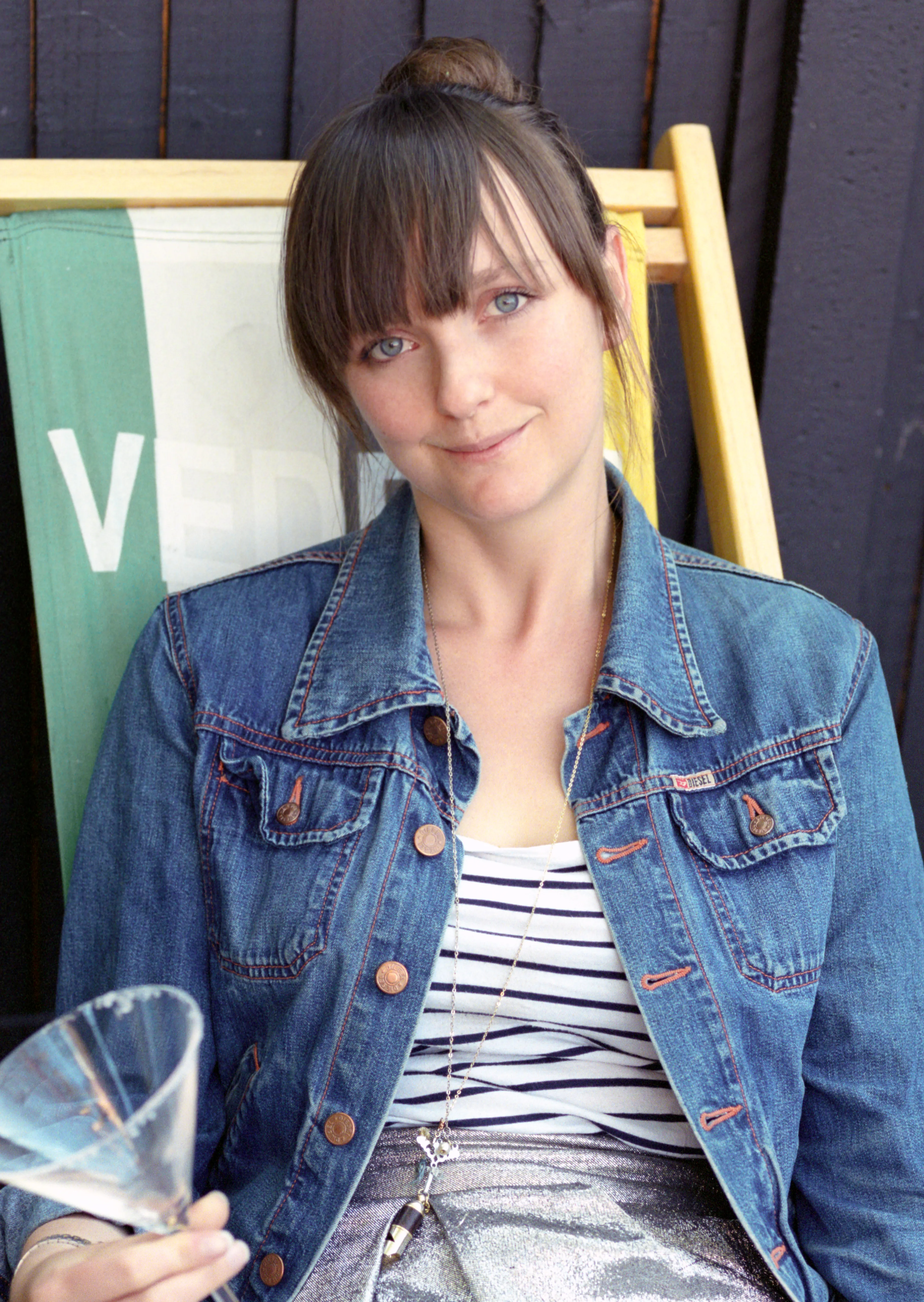Lauren seated in a deck chair in a denim jacket and a striped top, holding a cocktail glass and smiling slightly at the camera.