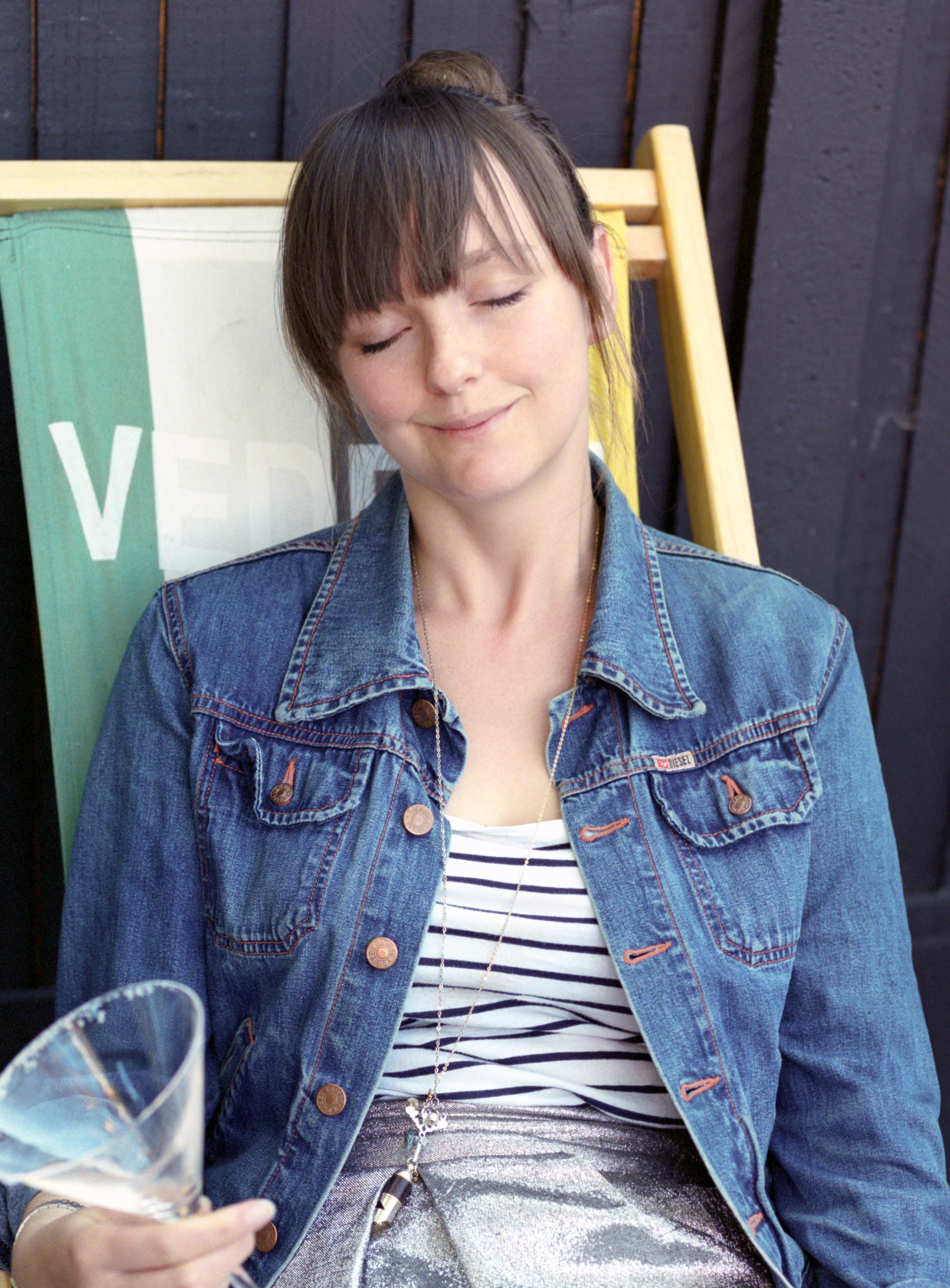 Lauren seated in a deck chair in a denim jacket and a striped top, eyes closed and smiling faintly, holding a cocktail glass.