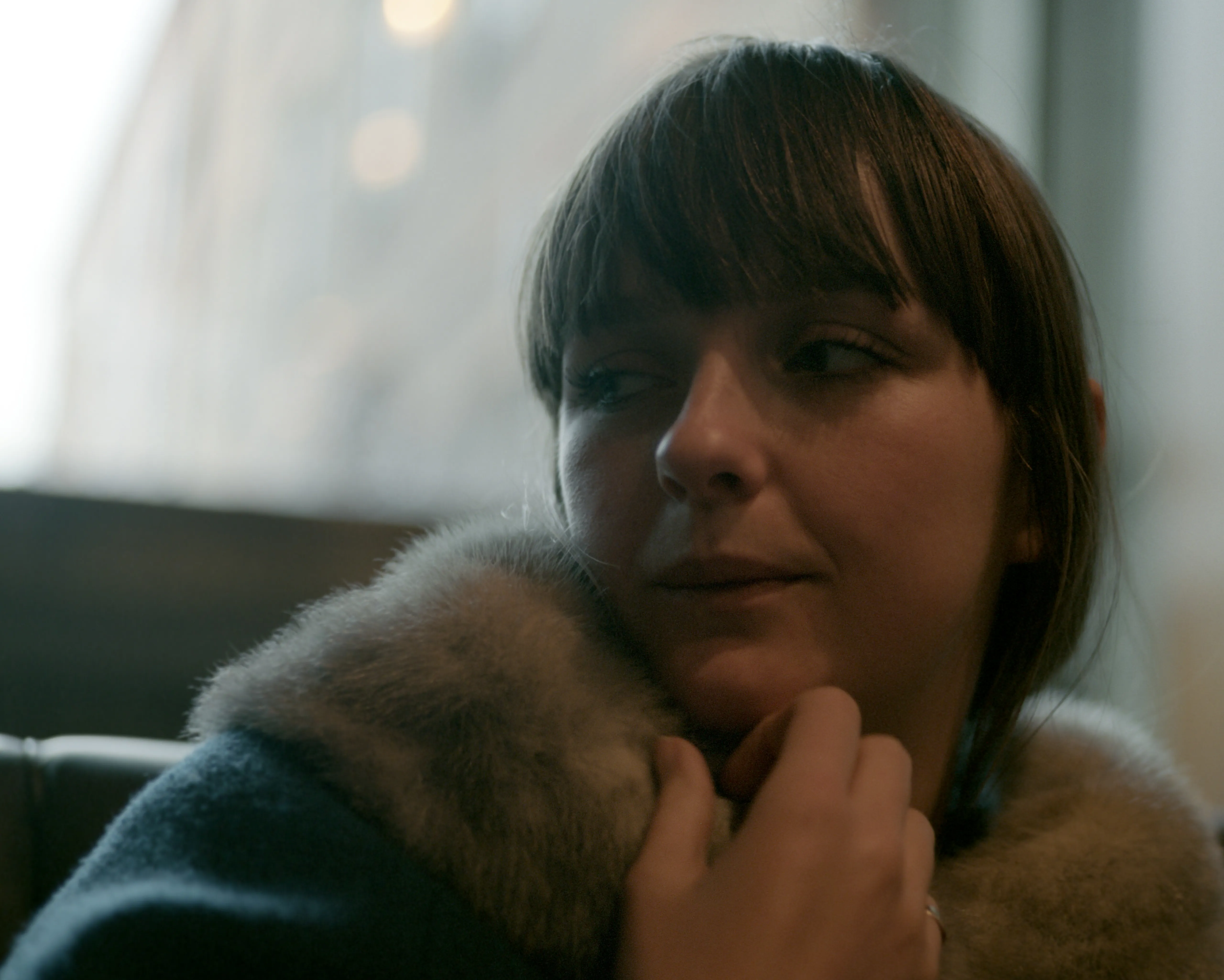 Close interior portrait of Lauren in a dark wool coat with a pale fur collar, fingers at her chin, looking off to the side; soft window light and a blurred bokeh behind her.