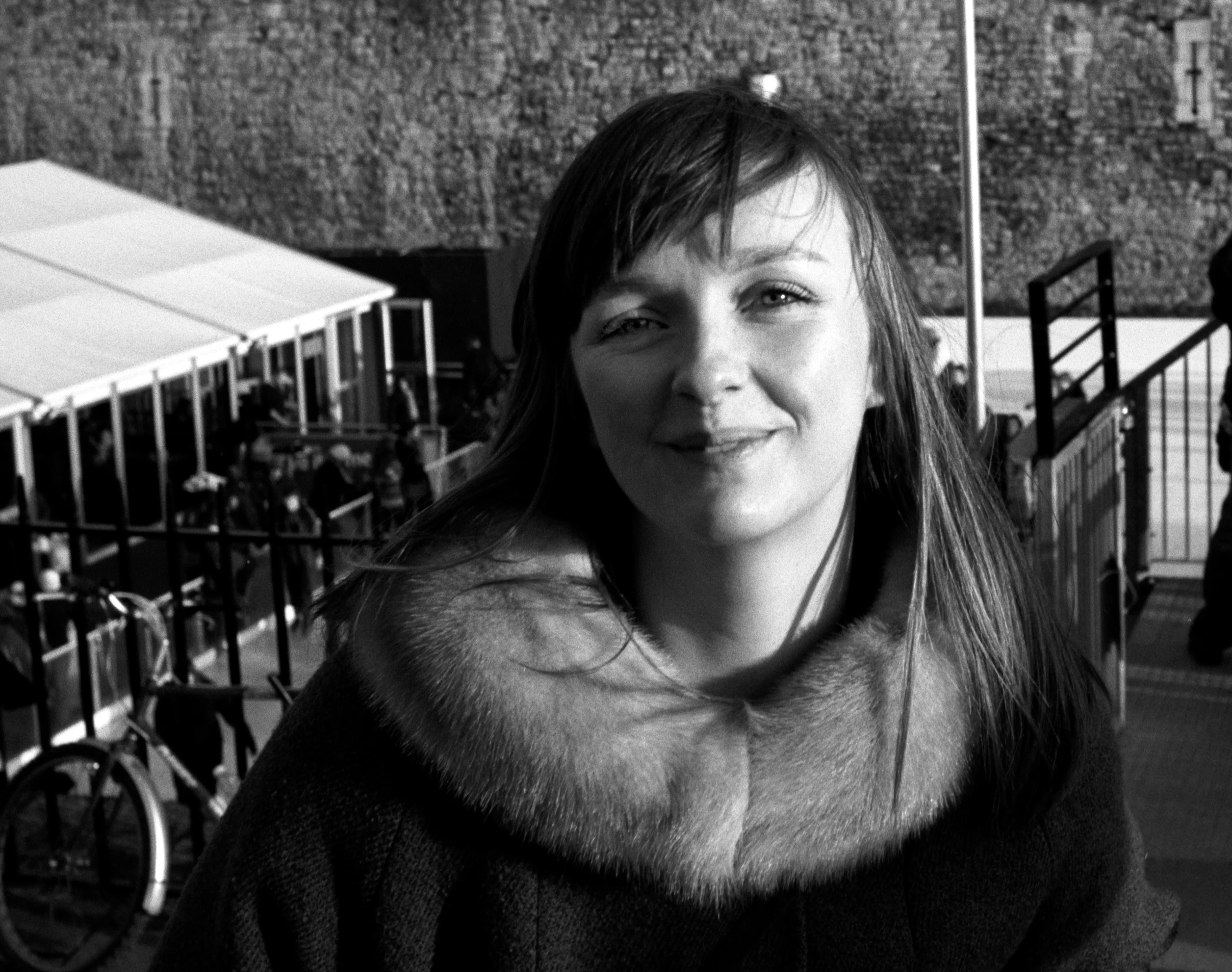 Lauren outside in a wool coat with a pale fur collar, hair loose across her shoulders, smiling faintly at the camera; a market stall and bicycles behind her. Black and white.