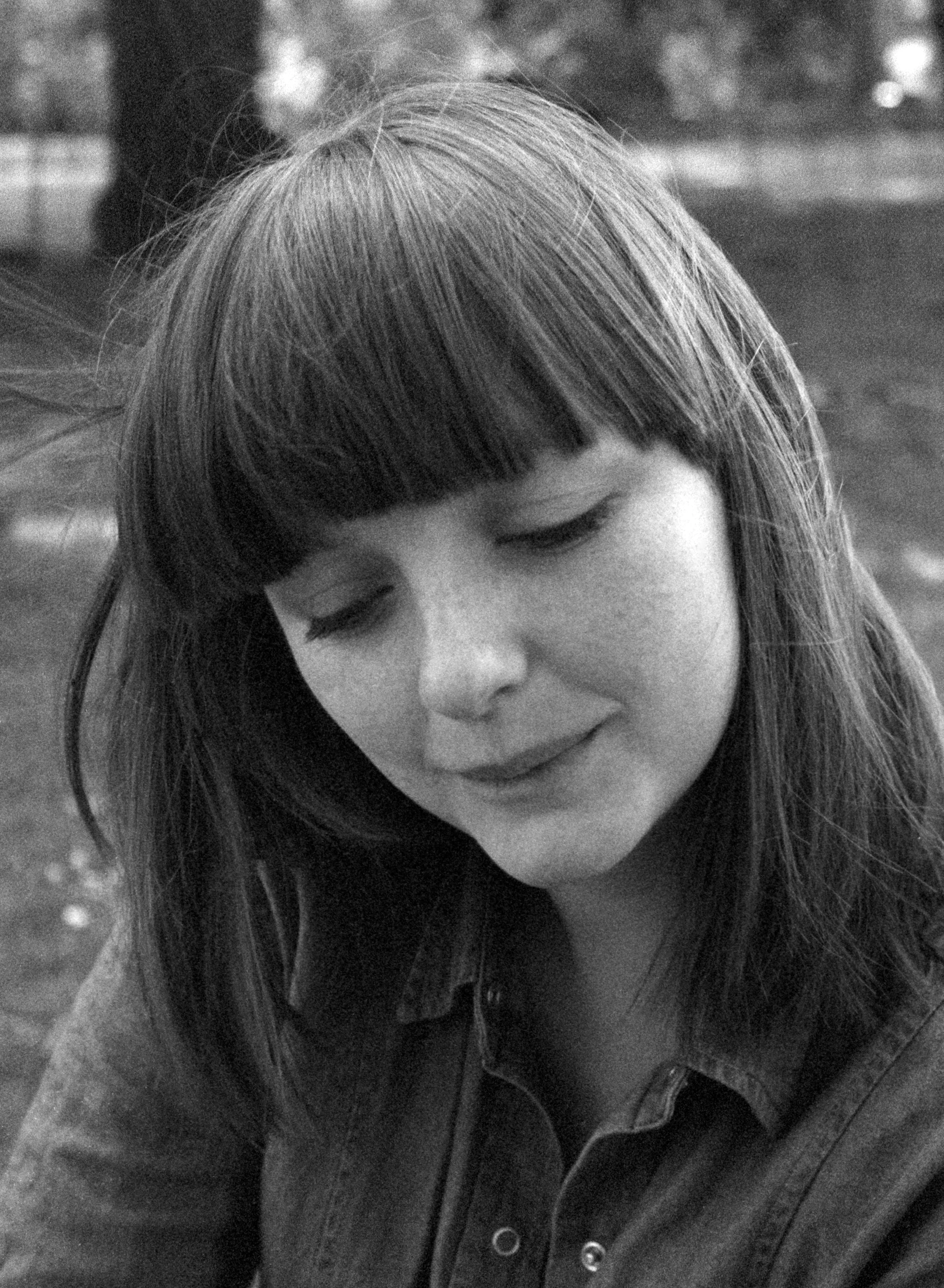Lauren with a dark fringe and loose hair, in a denim shirt, looking down with a faint smile in a park. Black and white.