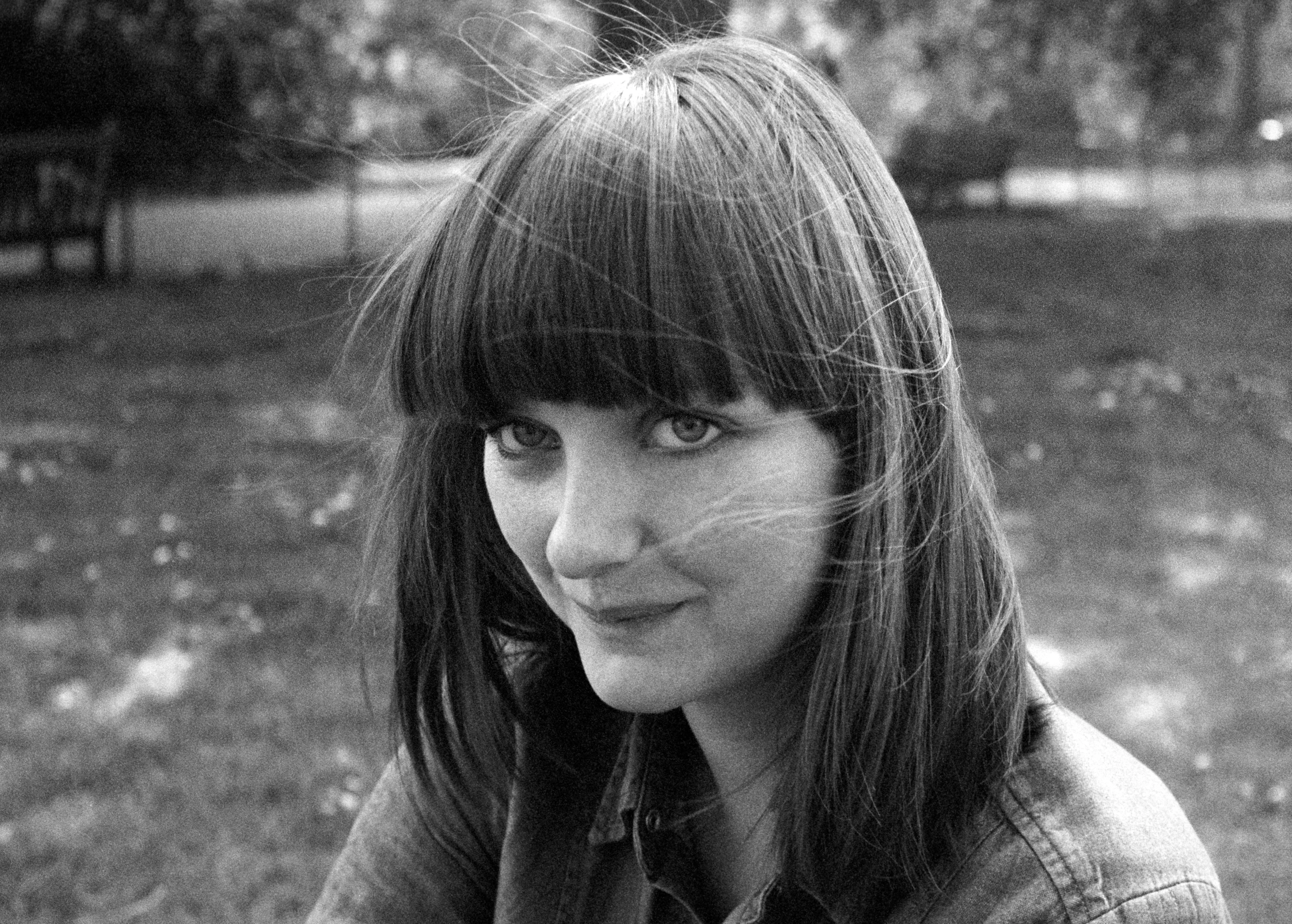 Lauren with a dark fringe and loose hair blowing across her face, in a denim shirt, looking directly at the camera in a park. Black and white.