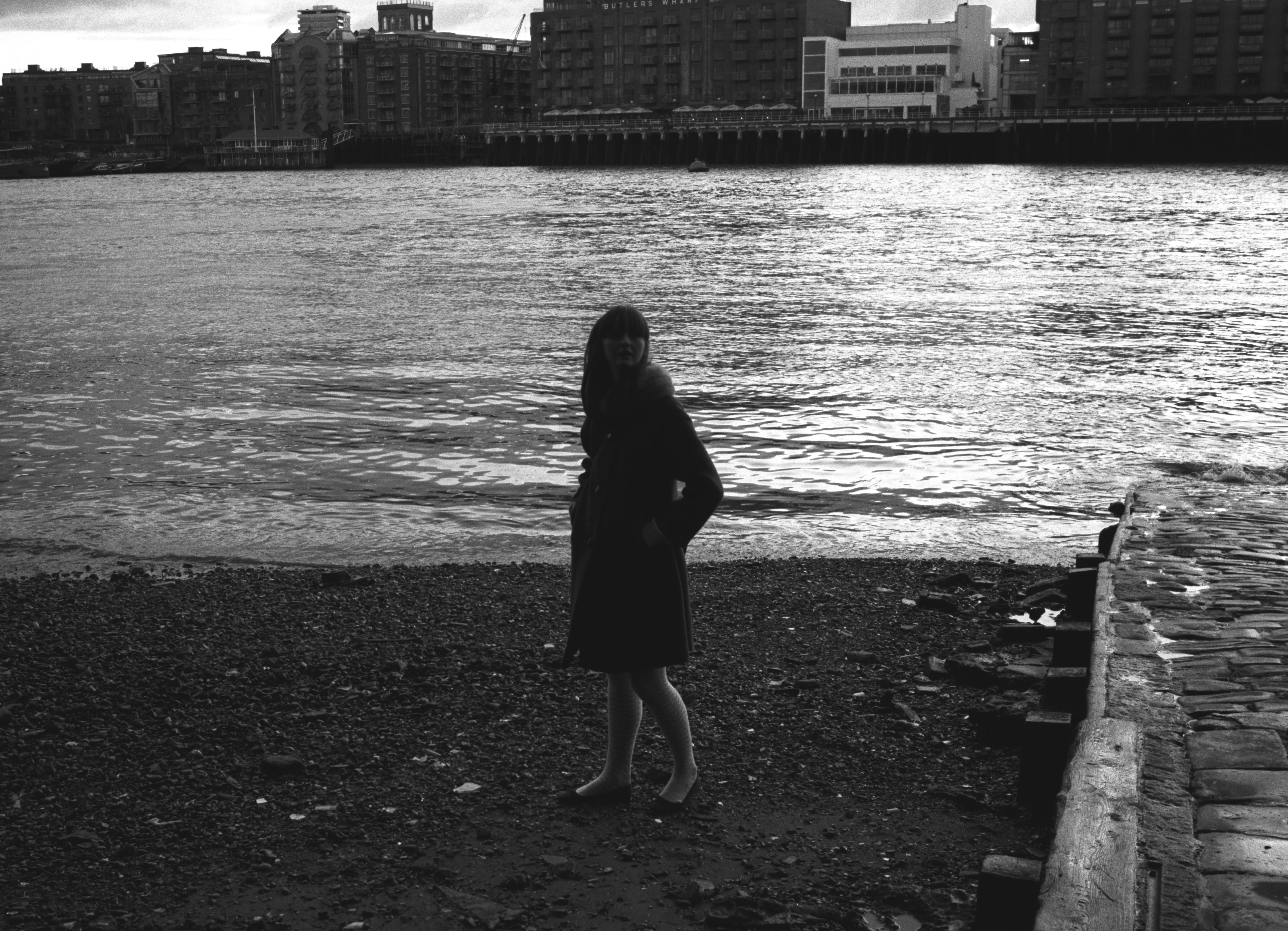Lauren on the Thames foreshore, standing barefoot on shingle in a dark dress, looking at the camera; the river and the City skyline beyond. Black and white.