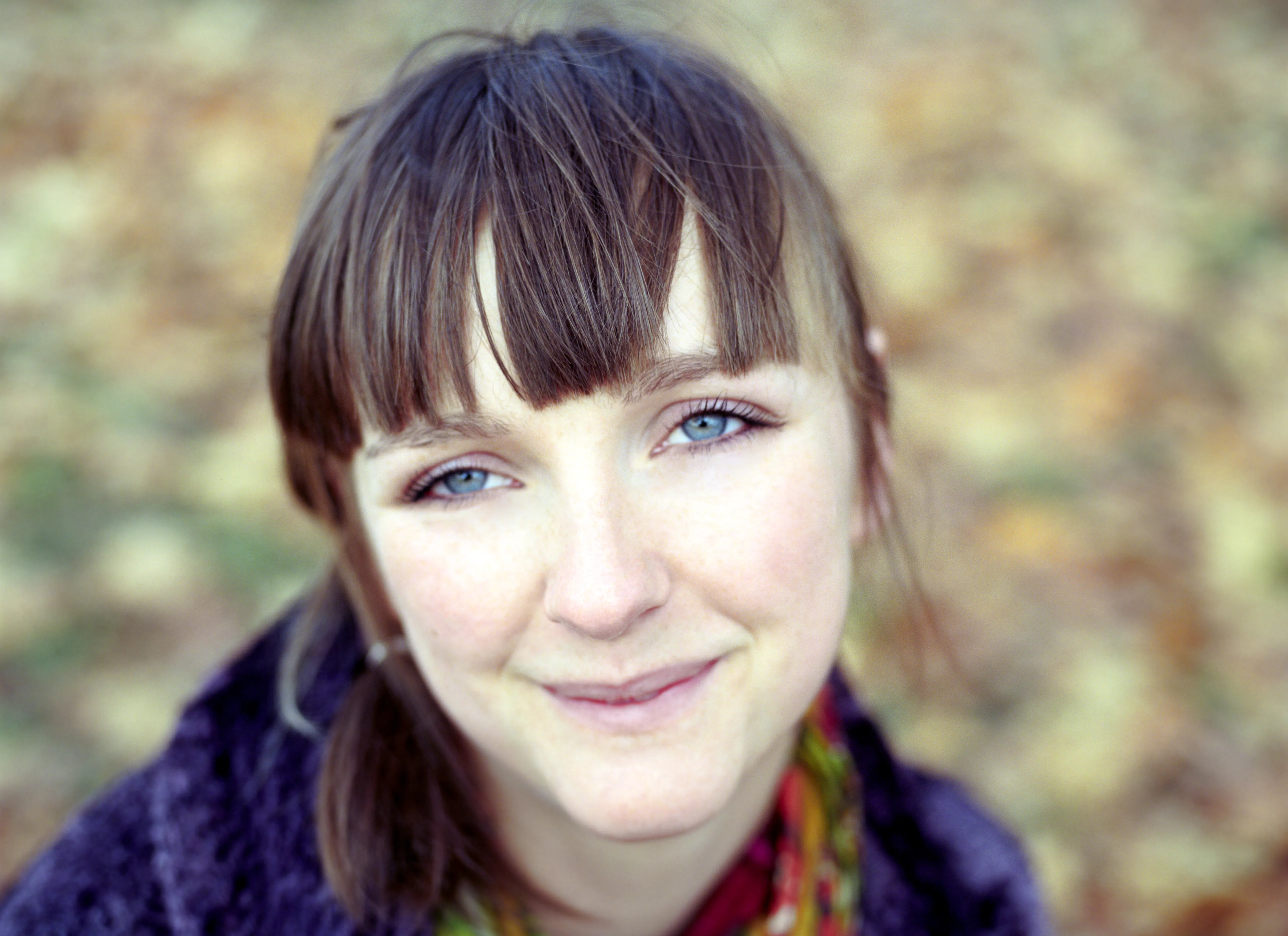 Lauren in a purple wool coat and a floral scarf, looking up at the camera with a calm closed-mouth smile, autumn woodland blurred behind her.