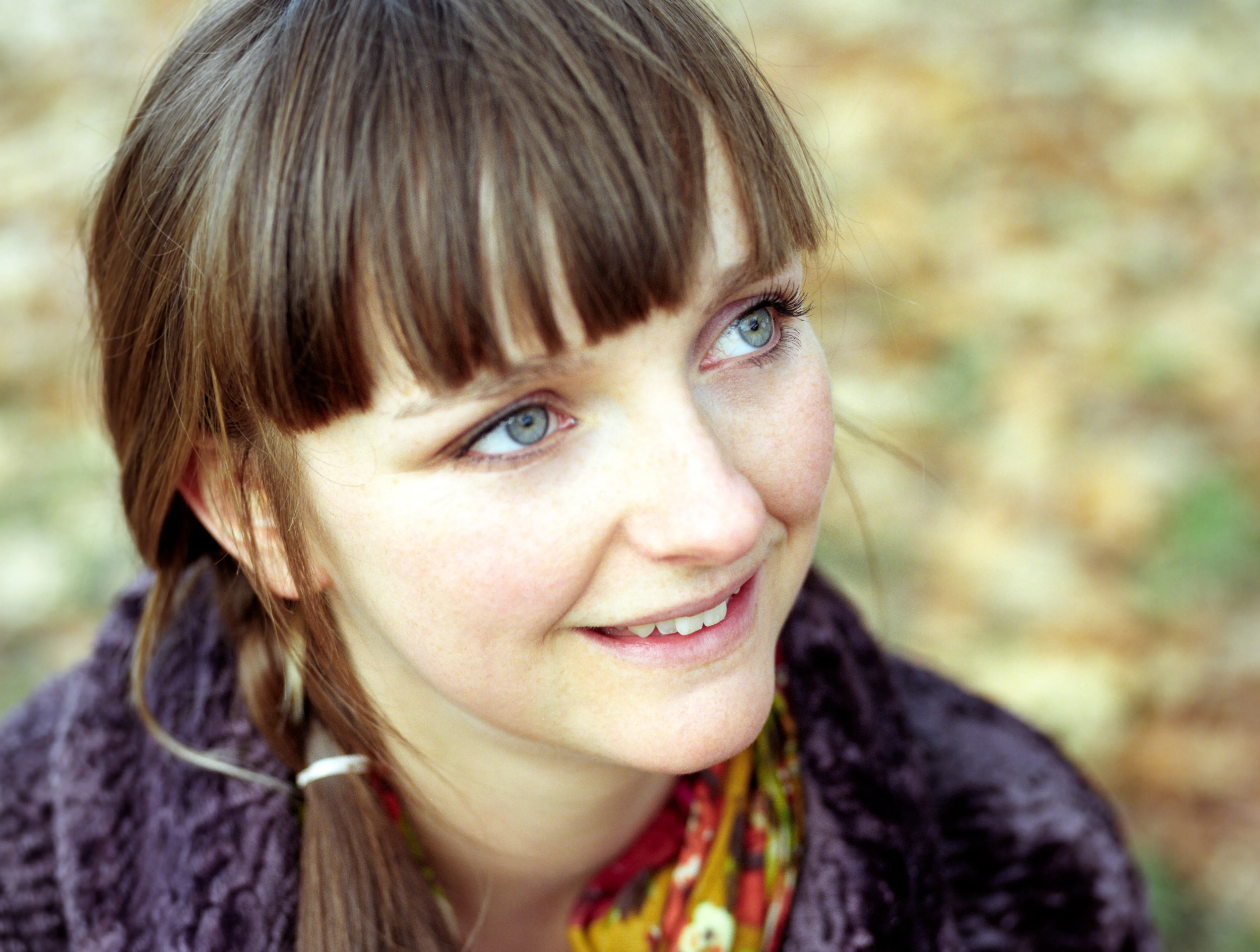 Lauren in a purple wool coat and a floral scarf, looking to one side with an open smile, autumn woodland blurred behind her.